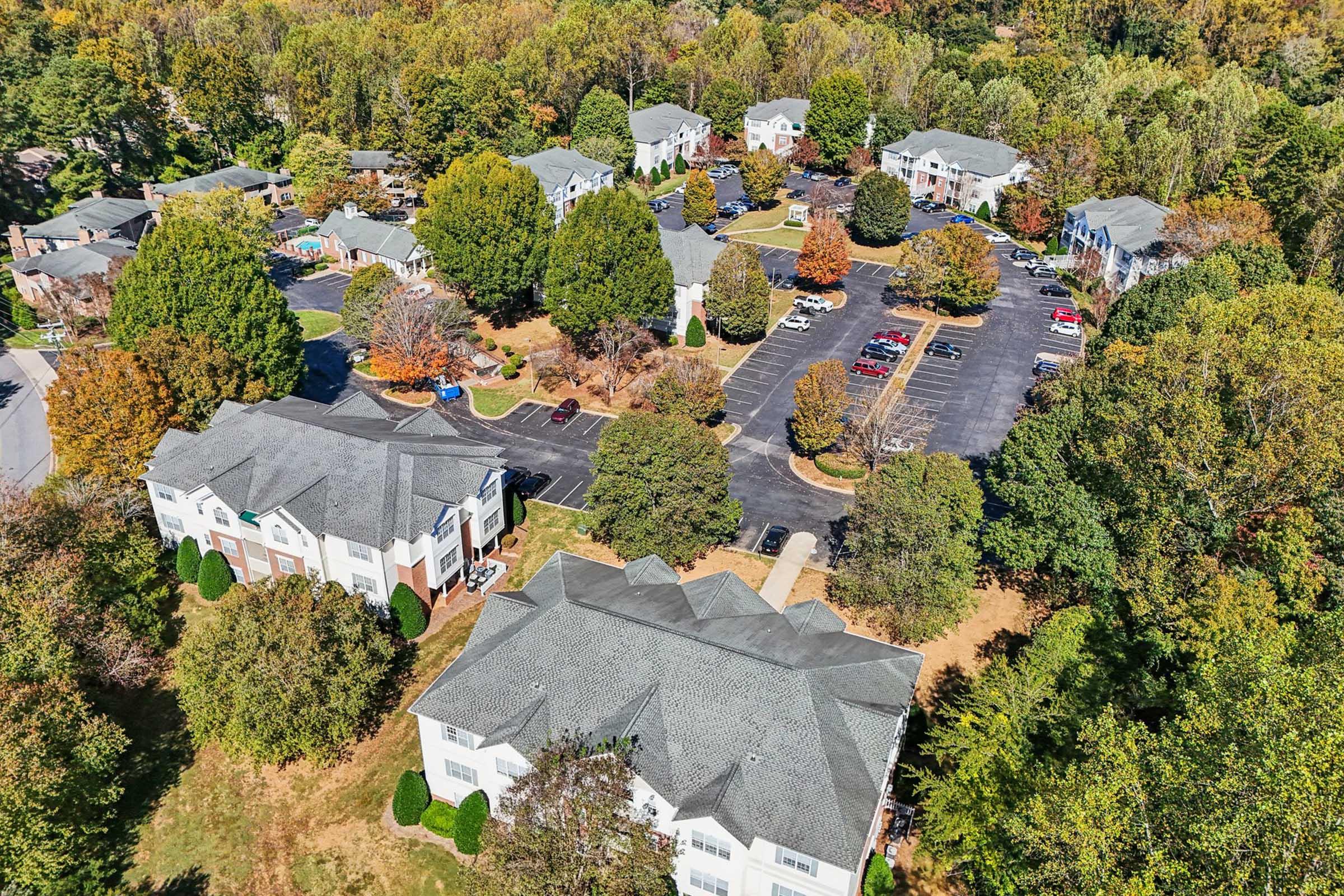 Aerial view of a residential area featuring multiple buildings surrounded by trees with vibrant autumn foliage. The parking lot is visible with several parked cars, and a small park or communal area is indicated by greenery in the middle of the complex.