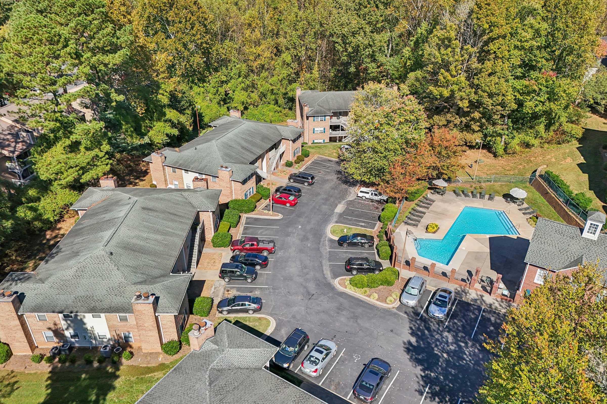 Aerial view of a multi-building residential complex surrounded by greenery. The image features a parking lot filled with cars, a swimming pool with lounge chairs, and landscaped areas, showcasing a serene living environment.
