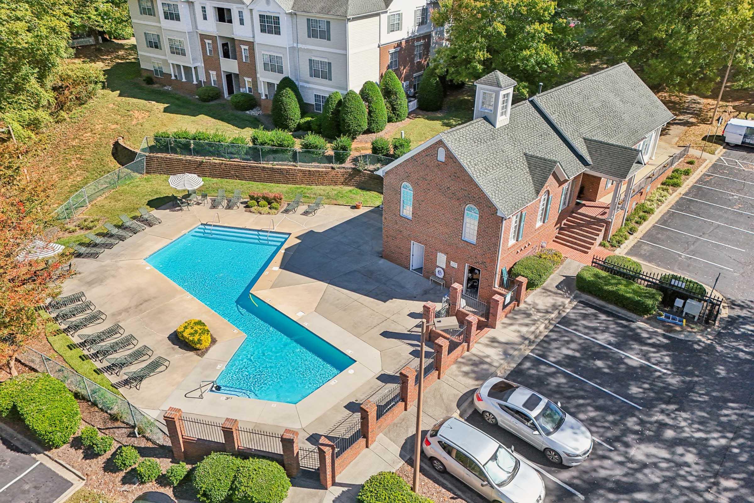 Aerial view of a swimming pool area featuring a rectangular pool surrounded by lounge chairs. There is a small building with a gable roof adjacent to the pool. The area is landscaped with hedges and trees, and there are parked cars in a nearby lot.