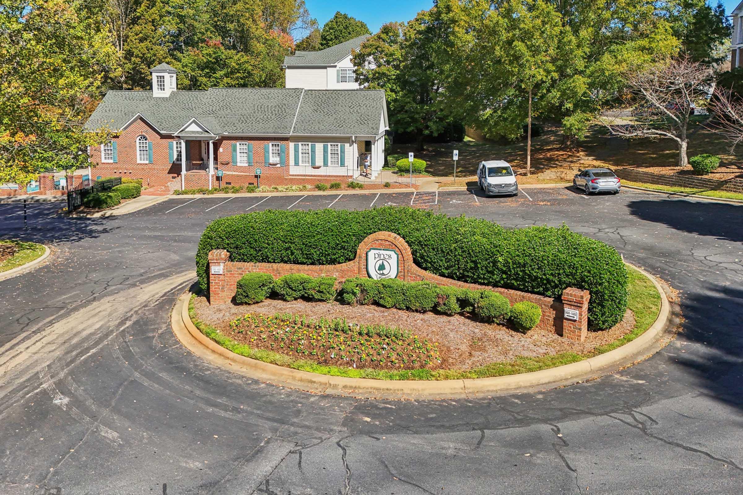 Aerial view of a neatly landscaped roundabout with a brick sign displaying the property name. Surrounding hedges and flowerbeds enhance the greenery. A few parked cars are visible in the background, with residential buildings partially seen among trees in a sunny, clear sky.