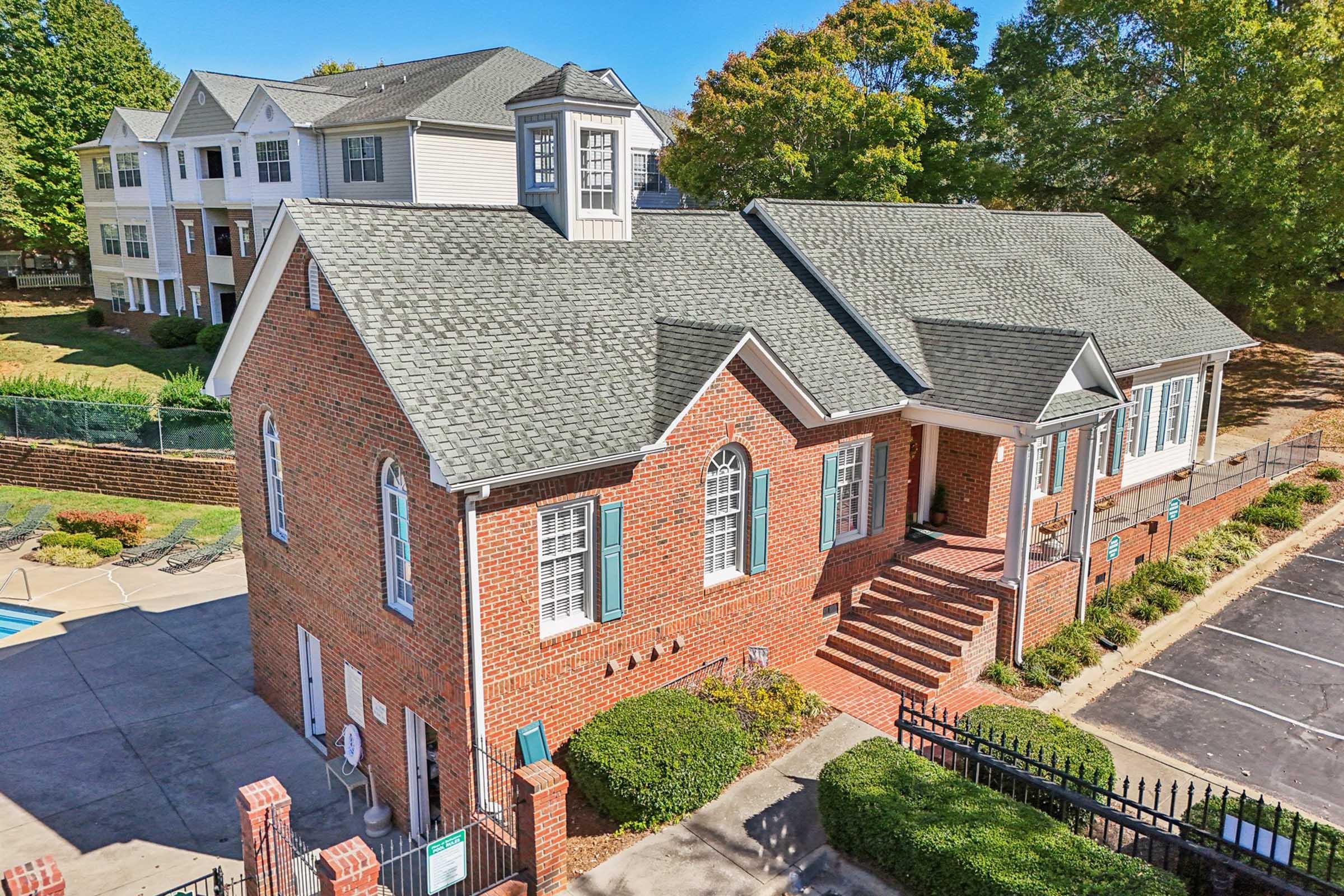 A brick building with a steep roof and decorative windows, surrounded by greenery. Adjacent to it, there is a parking area and a glimpse of a swimming pool. In the background, there are multi-story residential apartments. The scene is bathed in sunlight under a clear blue sky.