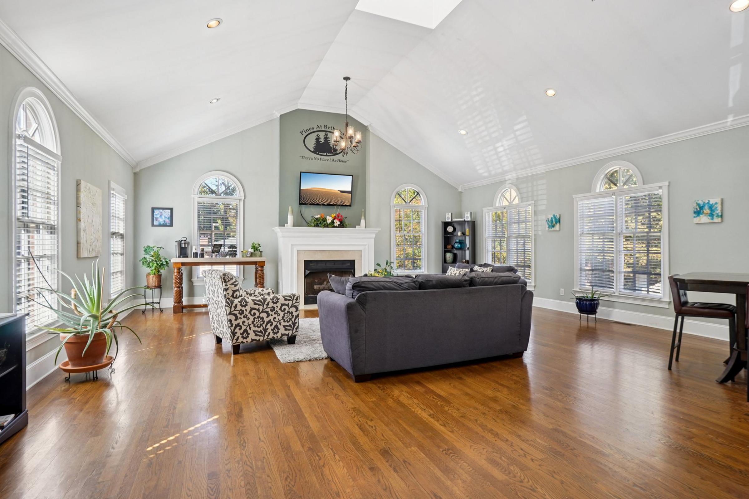 A spacious living room featuring a high ceiling with skylights, large windows, and hardwood flooring. It includes a cozy seating area with a dark sofa and a patterned chair, a fireplace, a television mounted above the mantel, and a small dining table in the corner. Decorative plants and artwork enhance the inviting atmosphere.