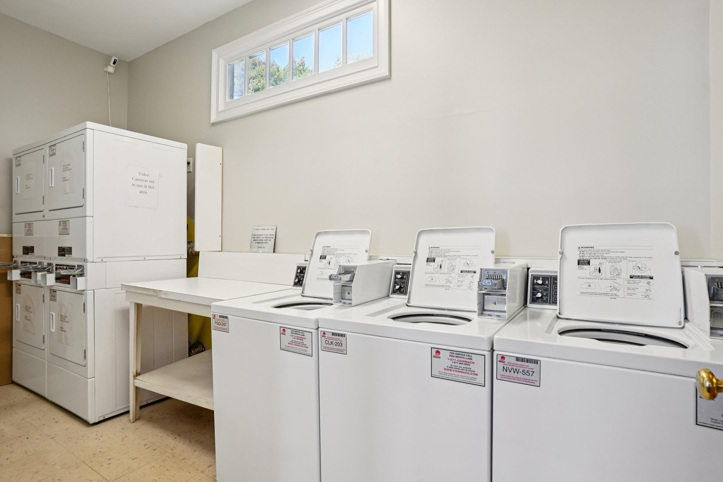 A laundry room featuring multiple white washing machines and dryers. The machines are arranged in a row, with a countertop and a window in the background providing natural light. Labels on the machines indicate model numbers and instructions. The setting is clean and organized, reflecting a functional space for laundry.