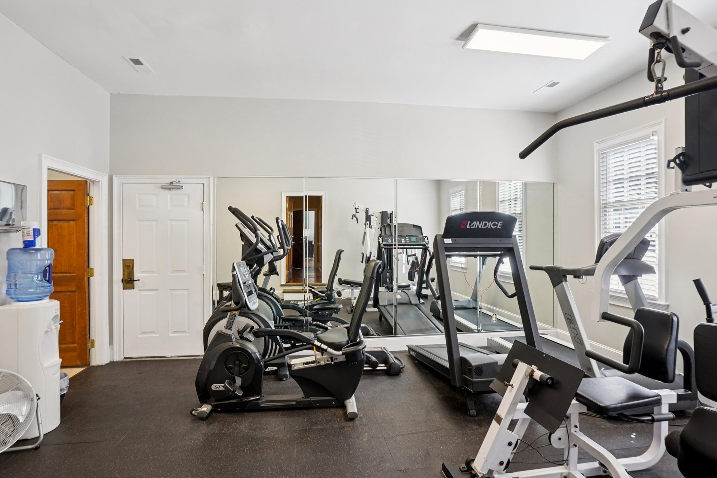 A well-equipped gym featuring exercise machines including treadmills, stationary bikes, and weight equipment. There are mirrors on the wall, a water cooler, and a fan, creating a spacious and functional workout environment. The room has bright lighting and a neutral color scheme.