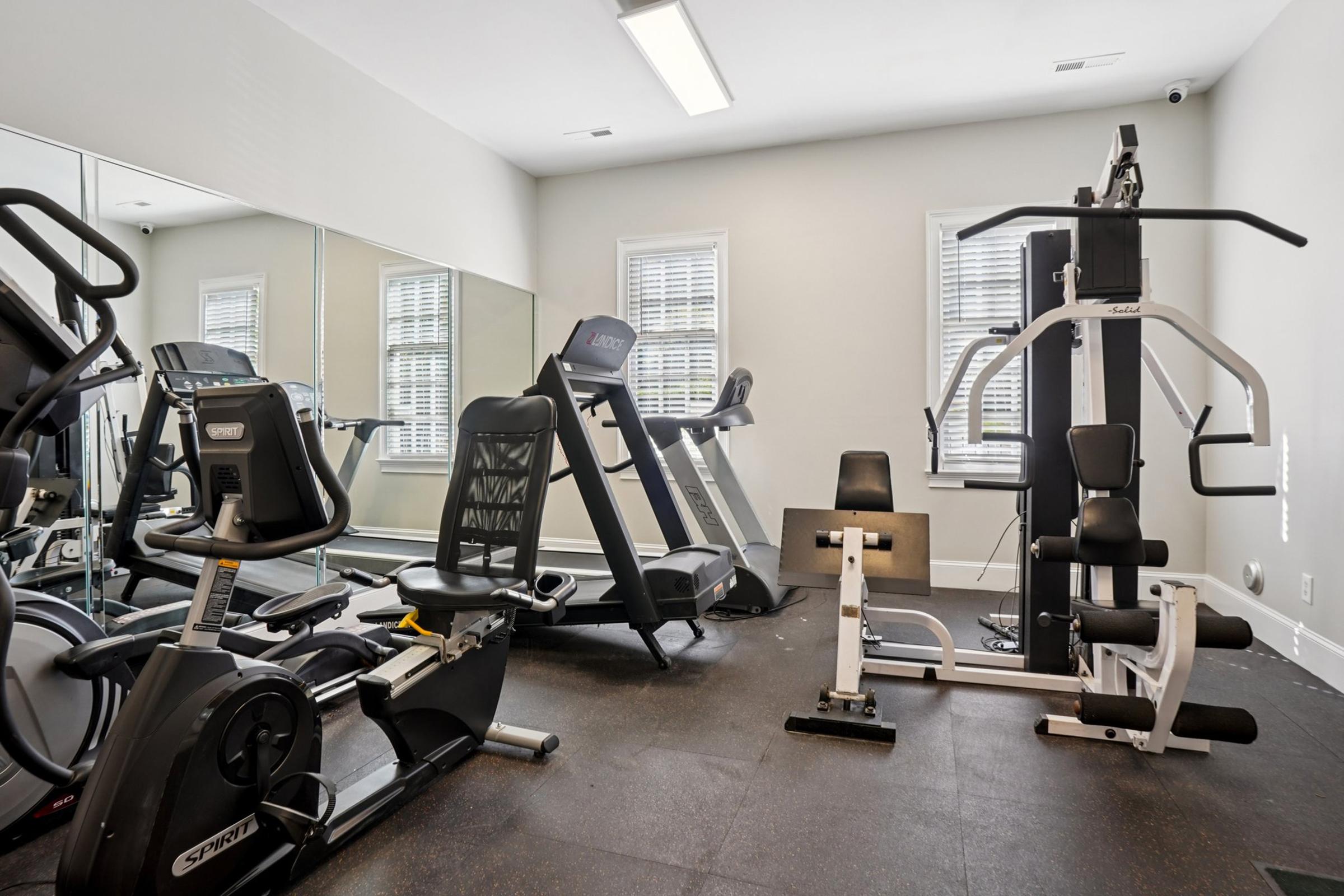 A fitness room featuring various exercise equipment, including a treadmill, stationary bike, weight machine, and free weights, with large mirrors and bright lighting. The space is designed for workouts and has a clean, modern aesthetic.