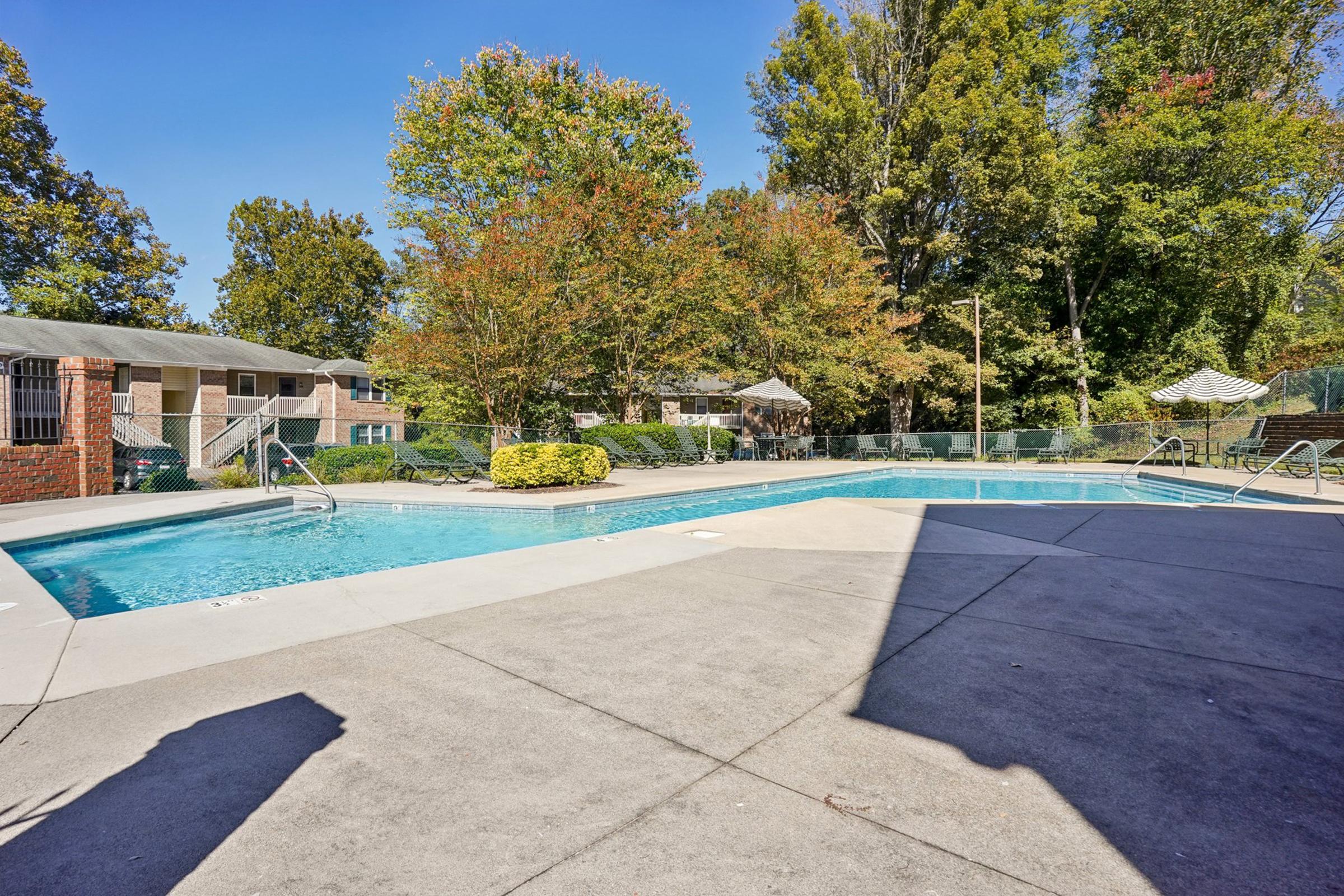 A sunny outdoor swimming pool area surrounded by trees and greenery. The pool features a shallow entry with steps, lounge chairs along the poolside, and a few shaded areas with umbrellas. Nearby, there are apartment buildings in the background, creating a relaxing atmosphere for residents.