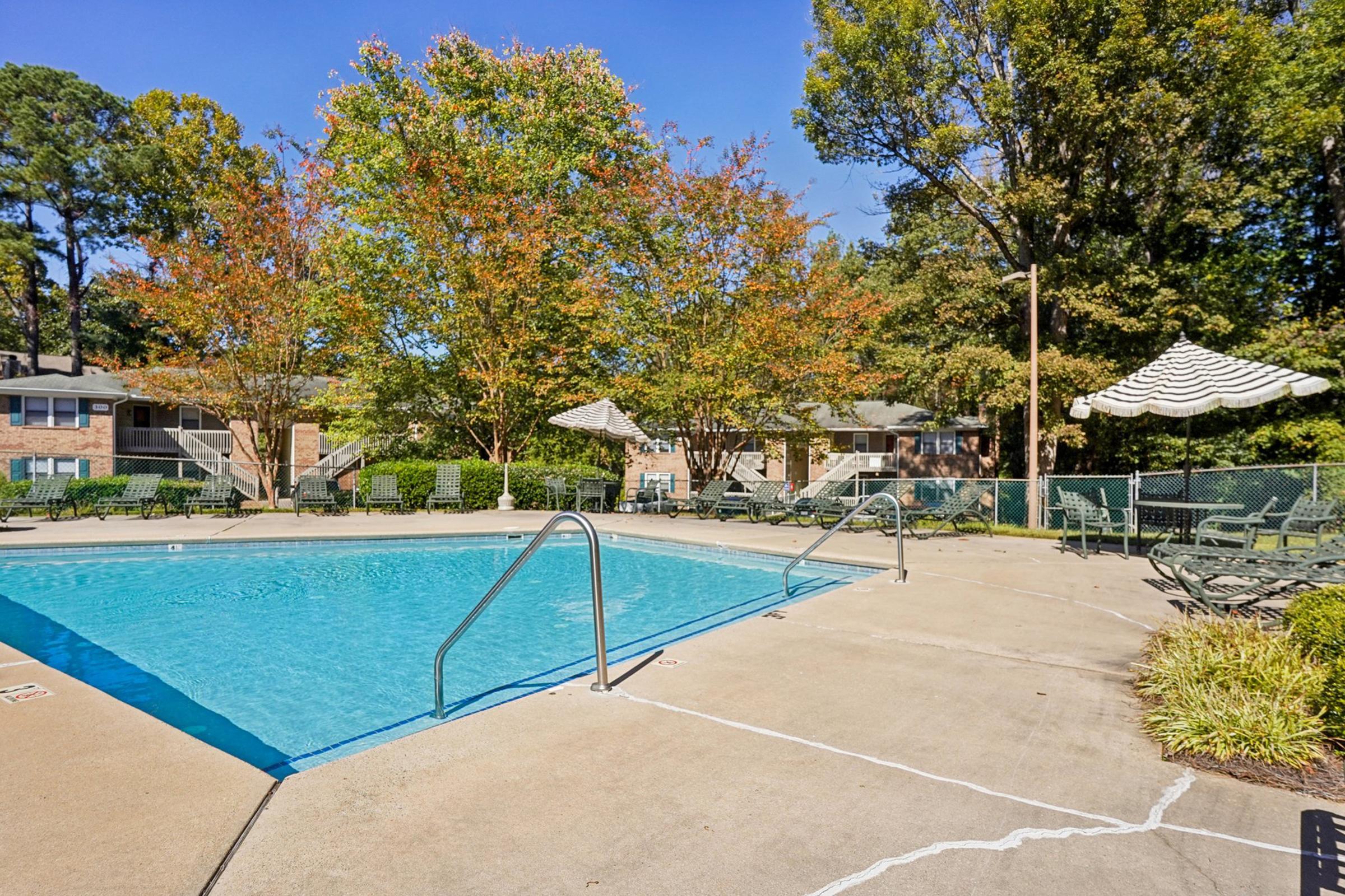 A clear swimming pool surrounded by a concrete deck, with lounge chairs arranged nearby. Colorful trees with autumn foliage provide a picturesque backdrop, and shaded umbrellas add to the inviting atmosphere. Nearby residential buildings are partially visible in the background under a bright blue sky.
