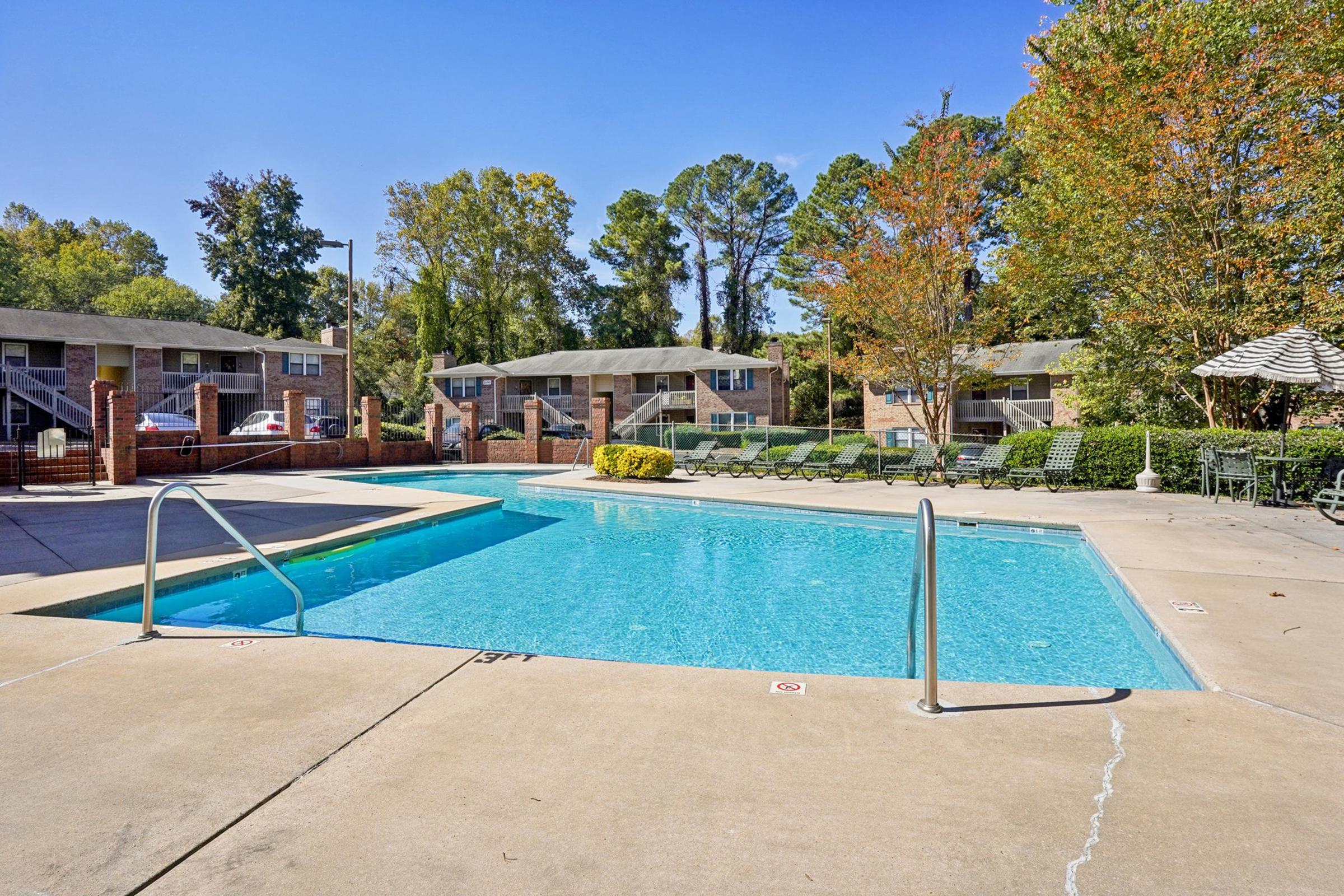 A clear blue swimming pool surrounded by a concrete patio and lounge chairs. In the background, there are several apartment buildings and lush trees with autumn foliage, indicating a pleasant outdoor setting. The pool area has a tranquil vibe, perfect for relaxation.