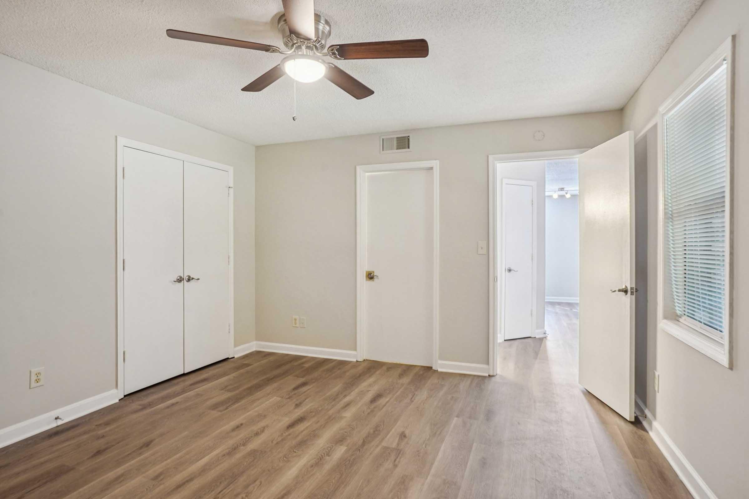 A well-lit interior room featuring light-colored walls and a ceiling fan. The floor is covered in light wood laminate, and there are two closed white doors on the left leading to closets. A doorway on the right leads to another room, with a window allowing natural light to enter.