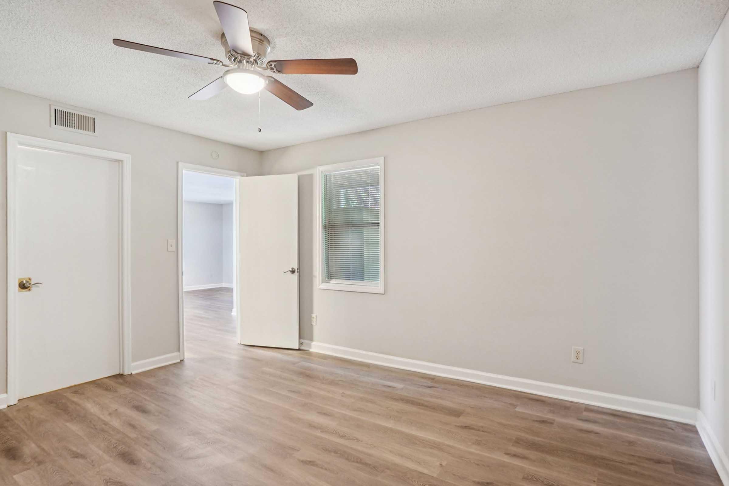 A spacious, well-lit room featuring a ceiling fan, light-colored walls, and laminate flooring. A door is partially open to another room, with a window allowing natural light. The overall atmosphere is bright and inviting, suitable for various uses such as a living space or office.