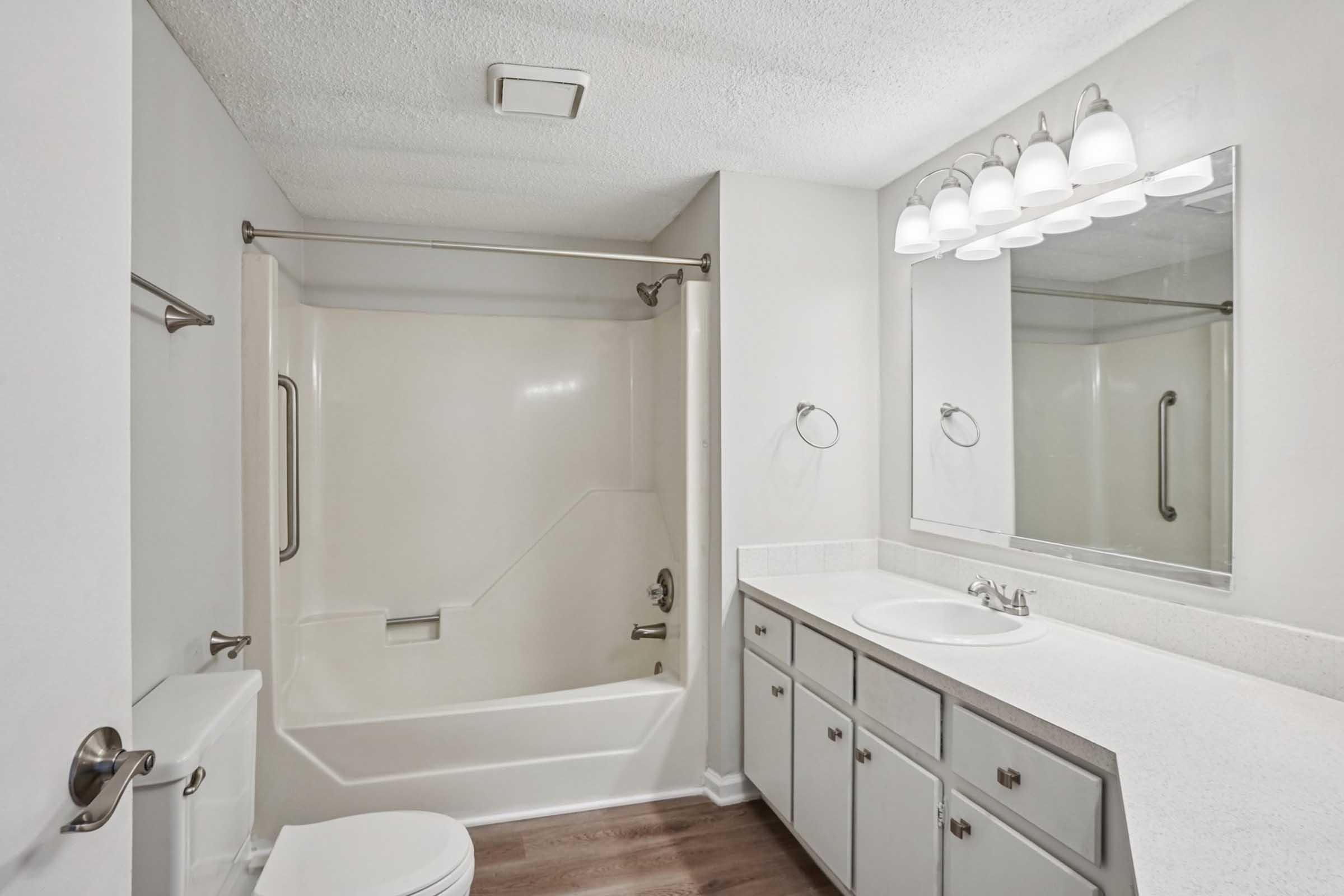 A clean, modern bathroom featuring a white shower and tub combination, a double sink vanity with a solid countertop, two mirrors above the sinks, and a neutral color scheme. The floor is finished with wood-like planks, and there are contemporary light fixtures above the mirror.
