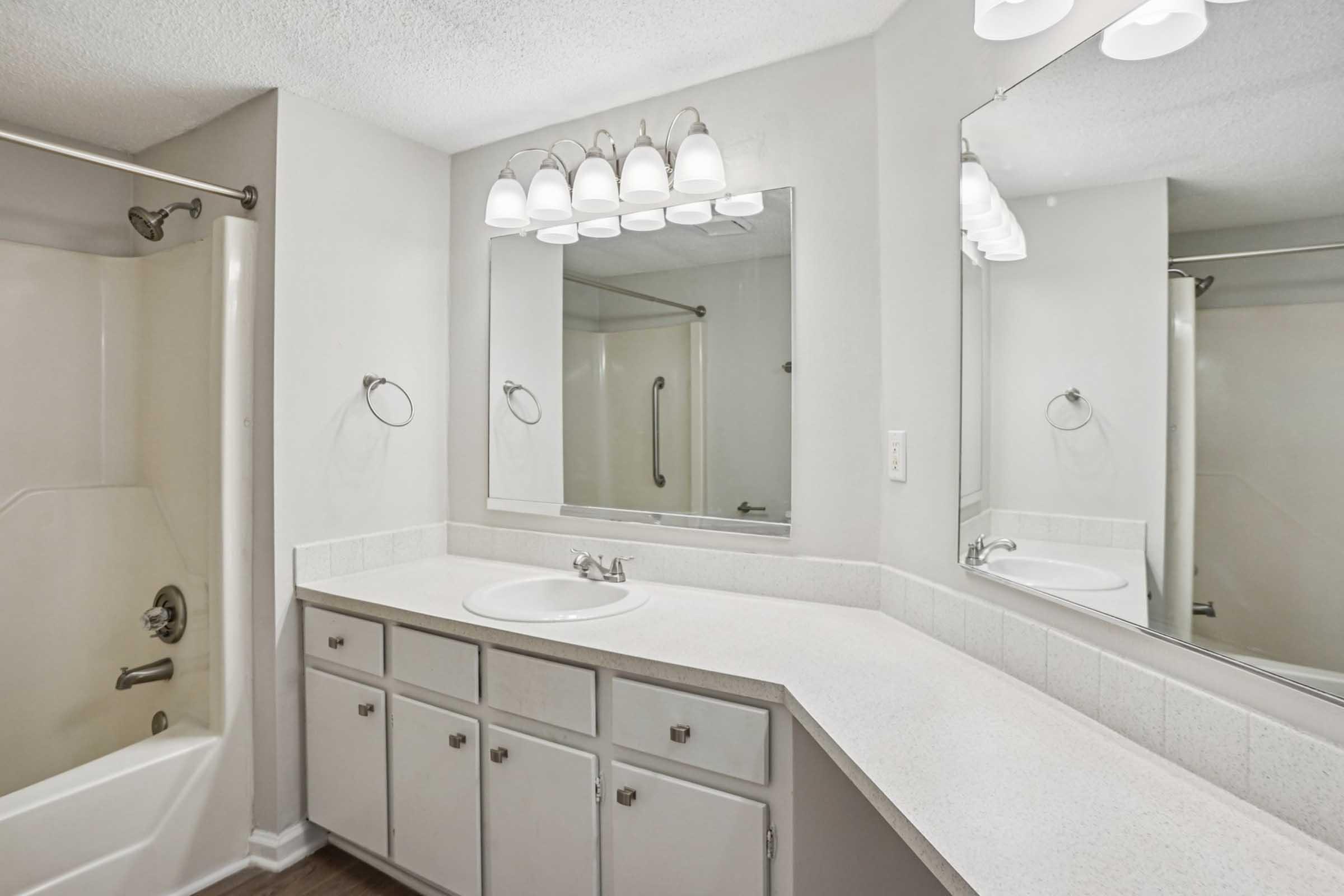 A modern bathroom featuring a white countertop with a sink and mirror, light fixtures above, and a bathtub and shower area. The walls are painted in a light color, and there are white cabinets below the countertop for storage. The overall design is clean and bright, providing a spacious feel.