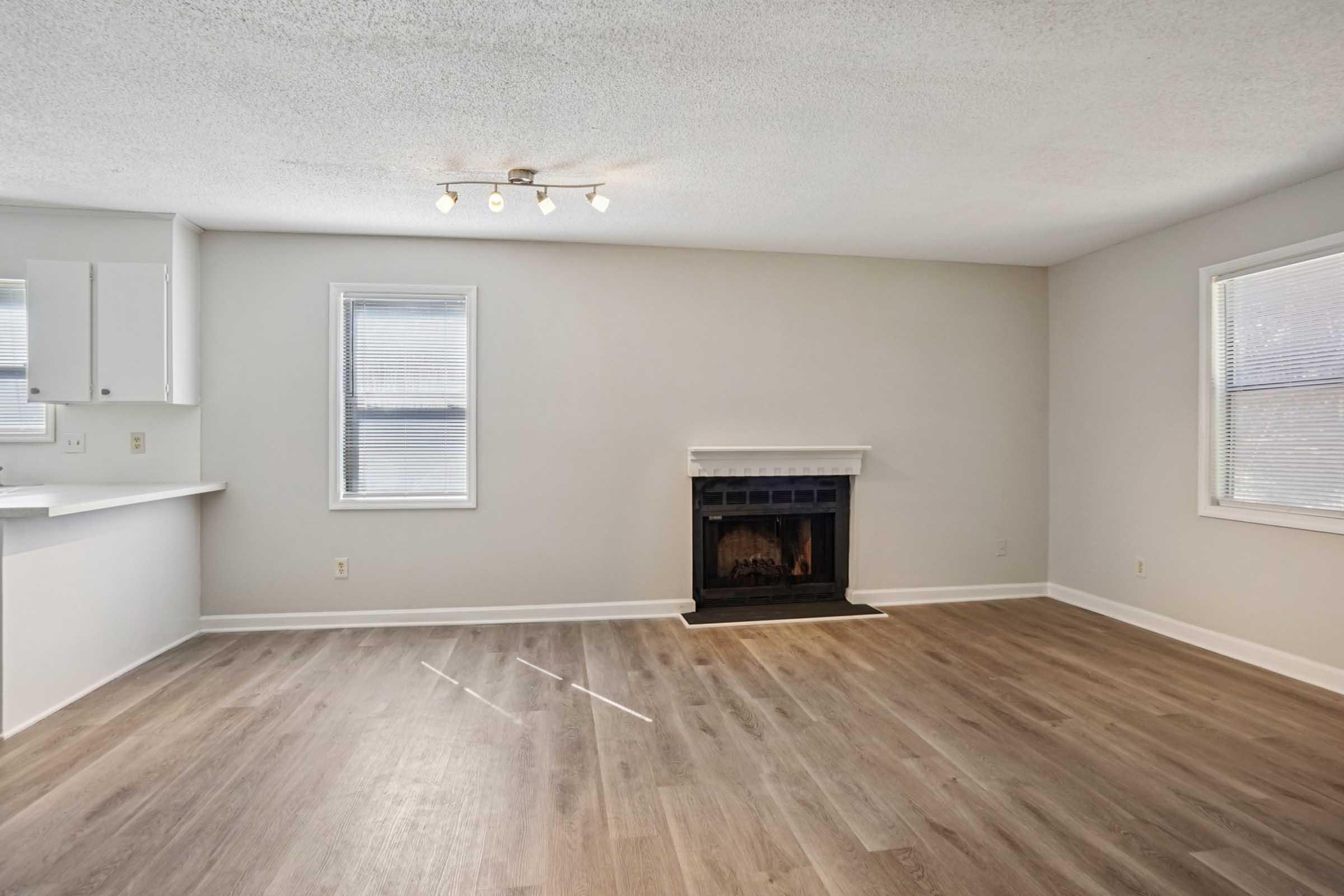 Bright and spacious living room with light-colored walls and hardwood flooring. A fireplace is centered against the far wall, flanked by two windows that provide natural light. The room has a modern feel and is partially connected to a kitchen area on the left.