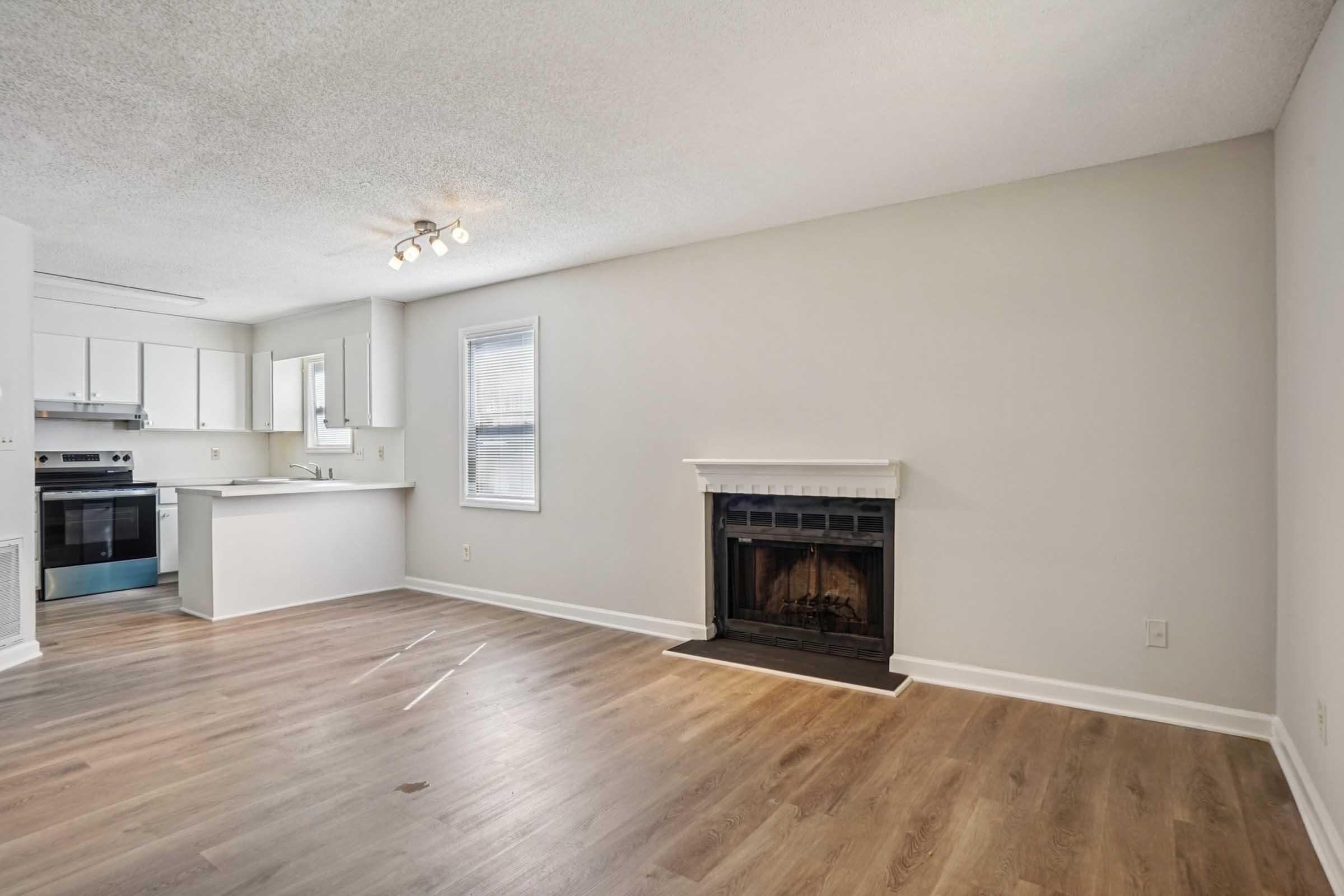 A bright and spacious living area showcasing light-colored walls and flooring. A traditional fireplace is the focal point on one wall, while a modern kitchen with white cabinets is partially visible in the background. Natural light flows through the windows, creating a welcoming atmosphere.