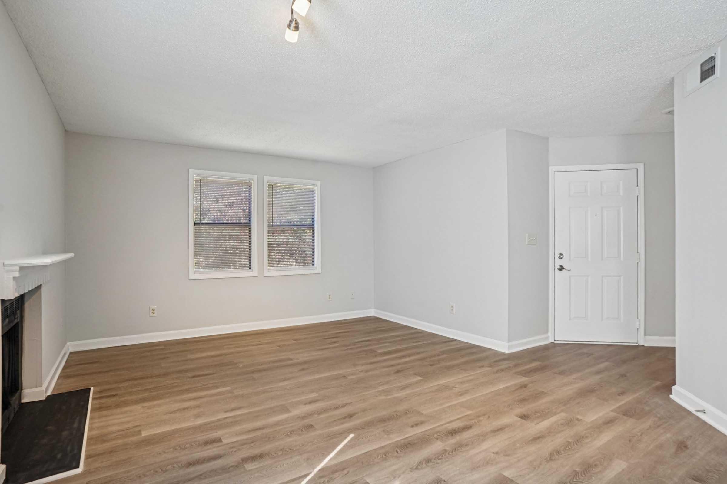 A spacious, empty living room with light brown wood flooring. Two windows allow natural light to filter in, and there is a white fireplace on one wall. The room is painted in a neutral gray tone, creating a calm and inviting atmosphere. A door leads to the outside, enhancing the feeling of openness.
