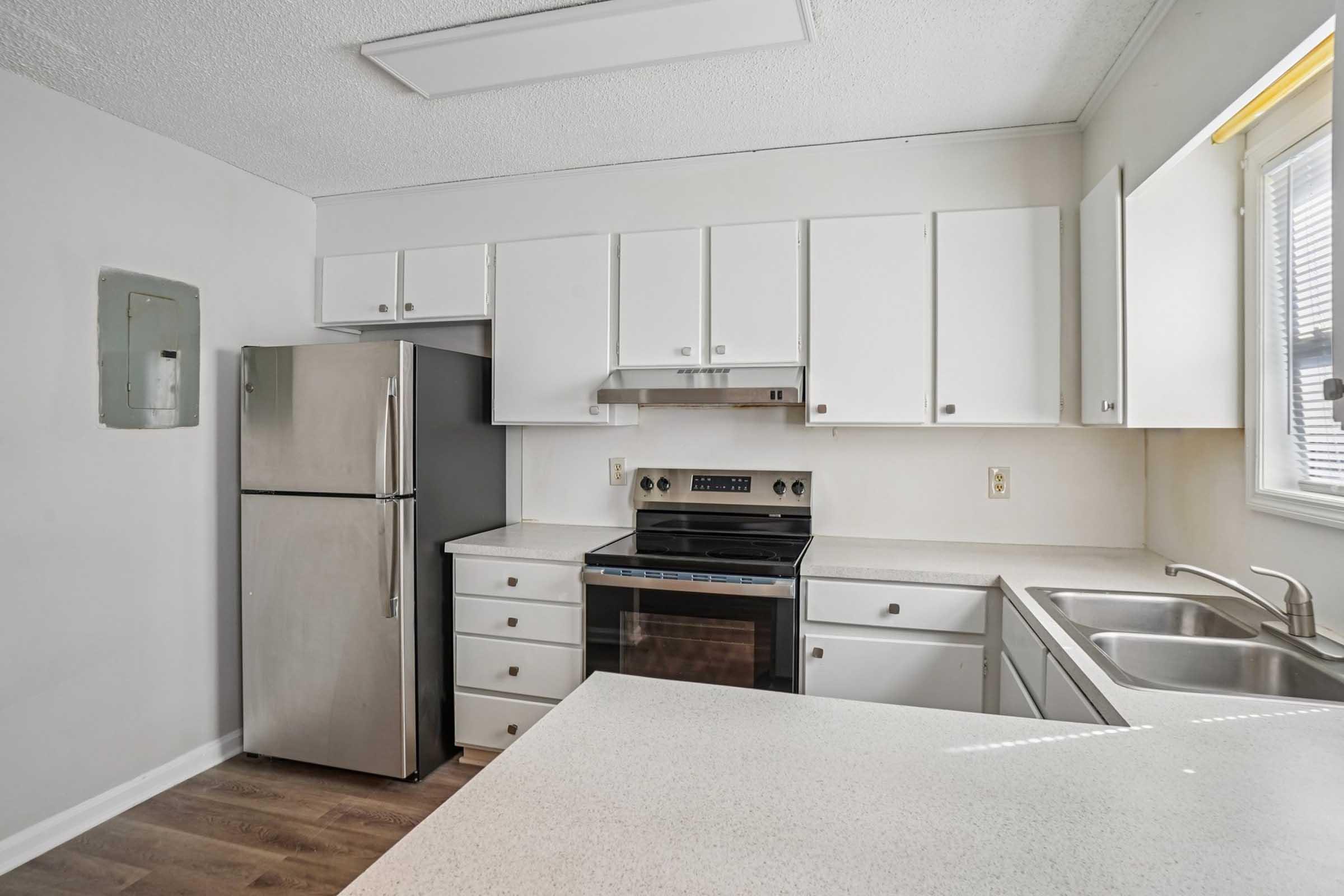 A modern kitchen featuring white cabinets, a stainless steel refrigerator, an electric stove, and a double sink. The countertop is light-colored, and there's natural light coming in from a nearby window. The space has a clean, contemporary design with ample storage.
