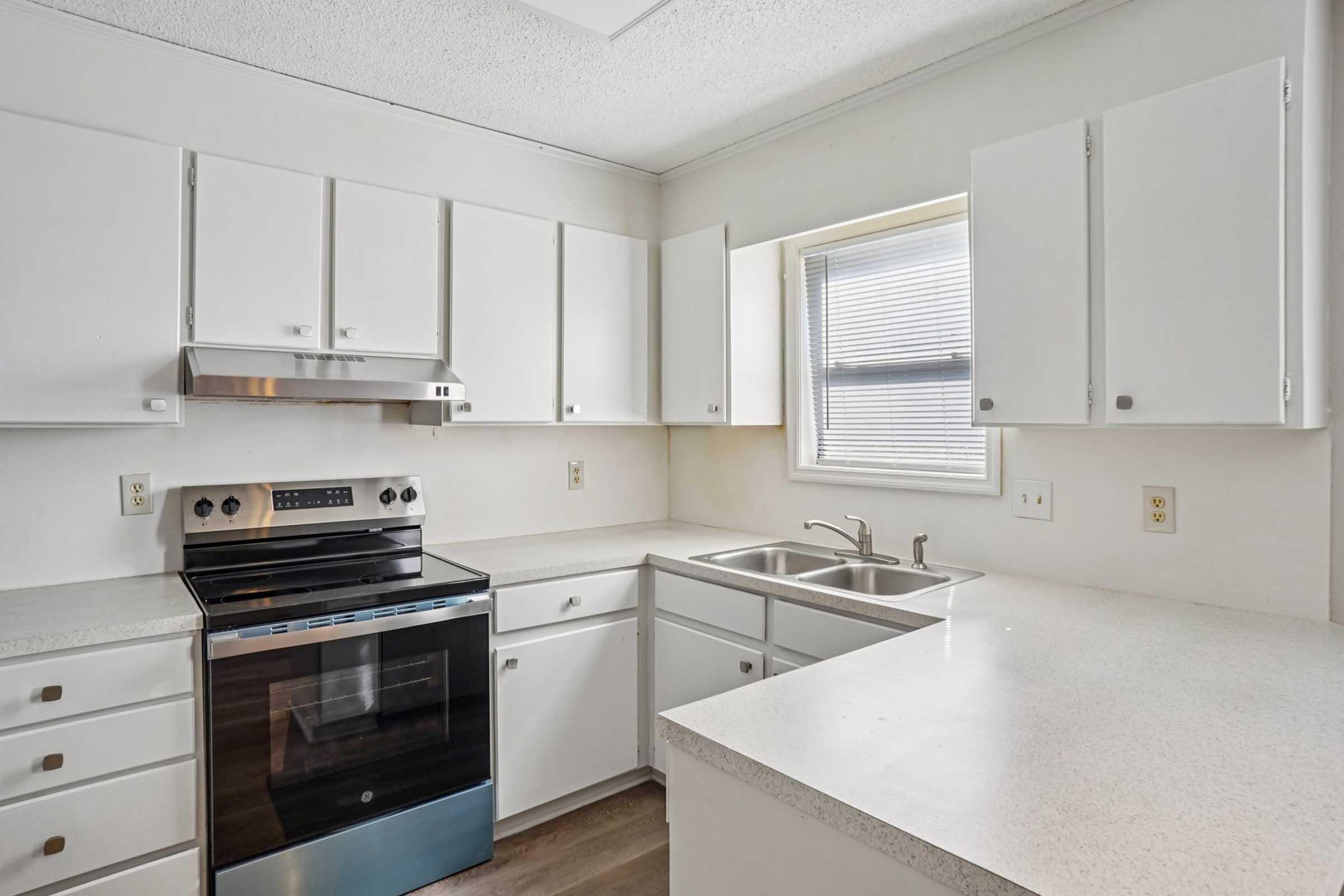 Bright, modern kitchen featuring white cabinetry, a stainless steel oven, and a dual-basin sink. Natural light streams in through a window above the sink, illuminating the clean countertops and sleek design.