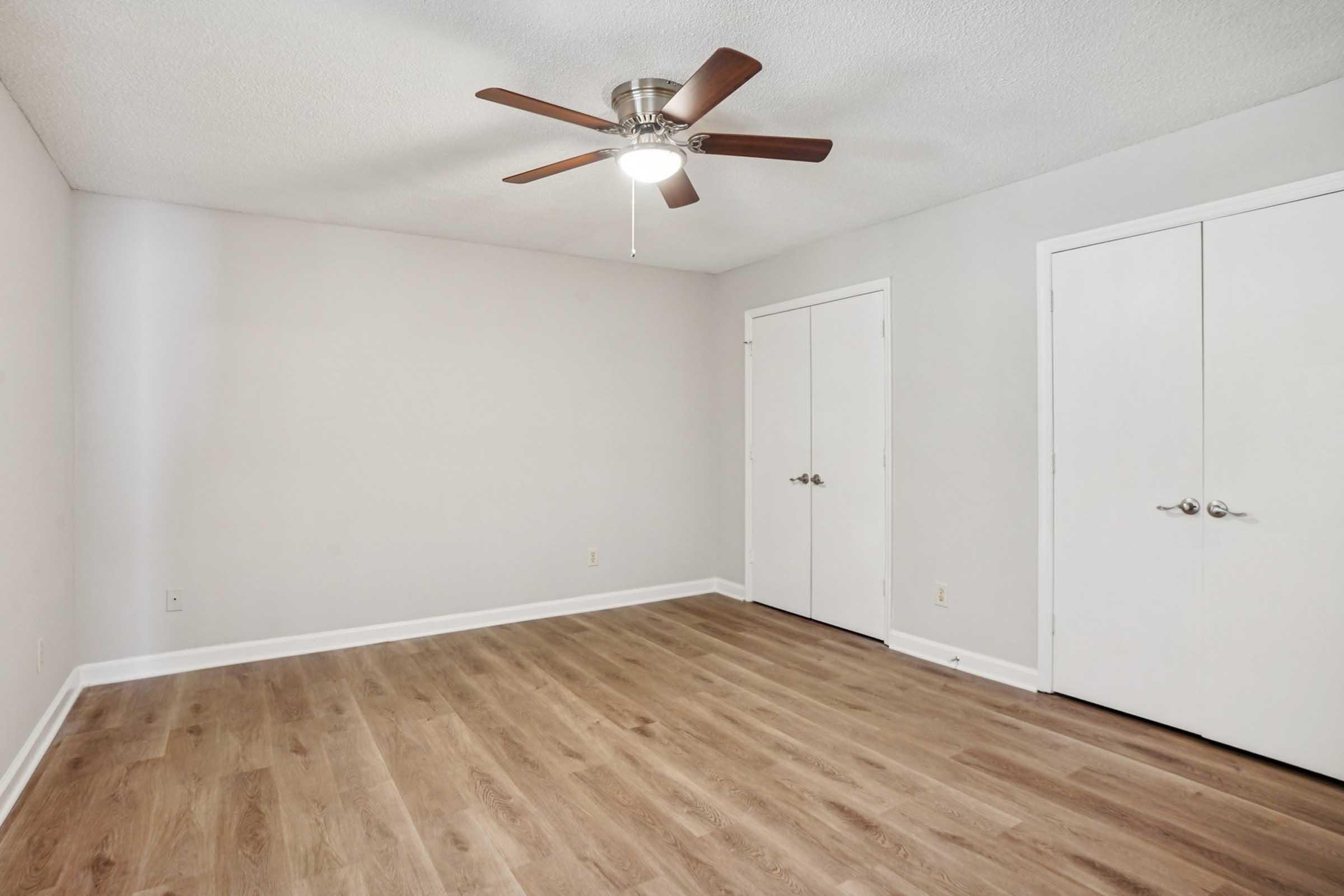 Spacious, empty bedroom featuring light gray walls, a ceiling fan with wooden blades, and laminate flooring. Two white double doors lead to storage or closets. Natural light fills the room, creating a bright and airy atmosphere, ideal for personal decoration and furniture arrangement.