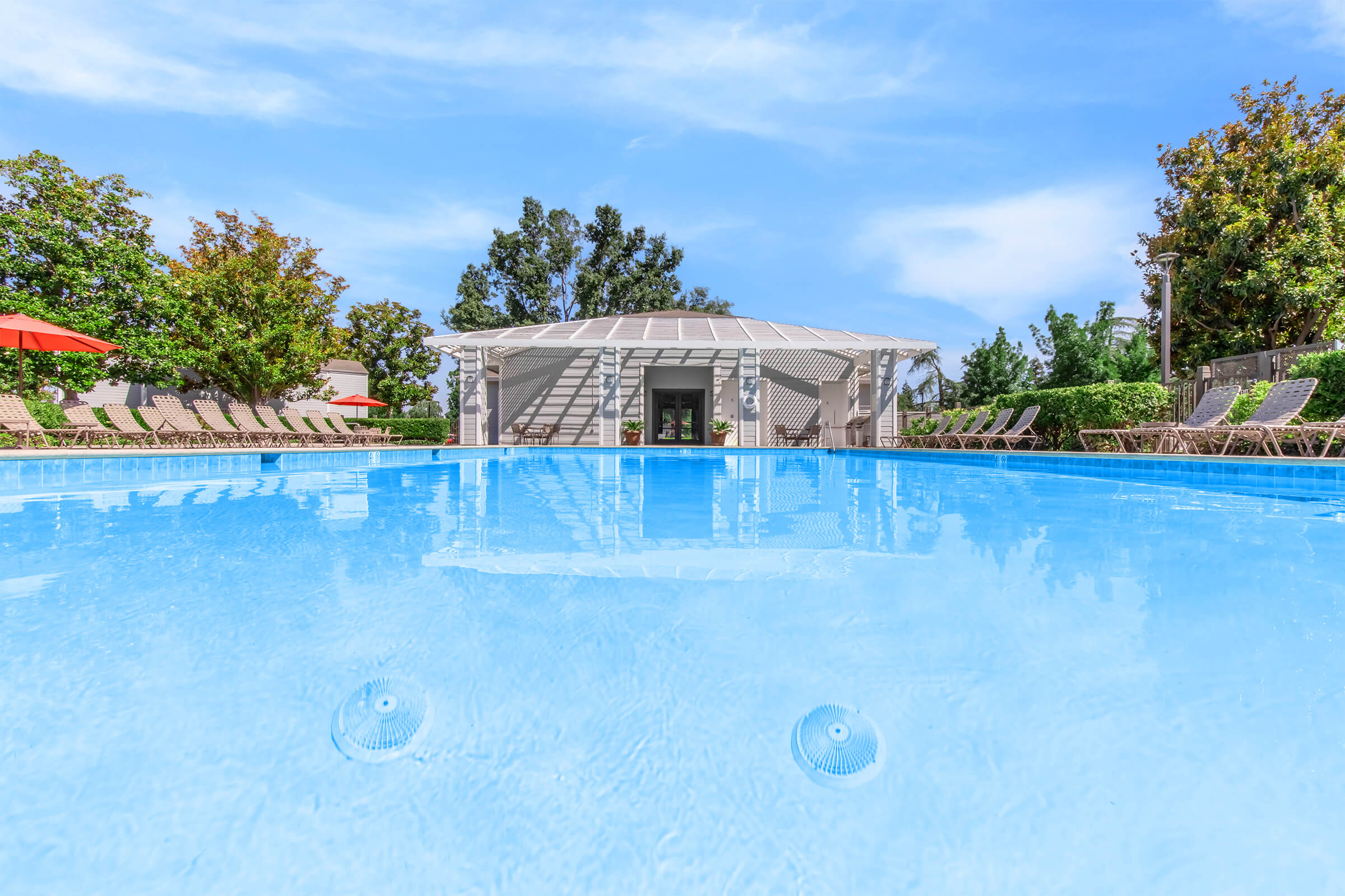 A tranquil swimming pool surrounded by lounge chairs and shaded areas, with a modern pavilion in the background. The clear blue water reflects the sky, creating a serene atmosphere, ideal for relaxation and leisure. Lush greenery adds to the picturesque setting.
