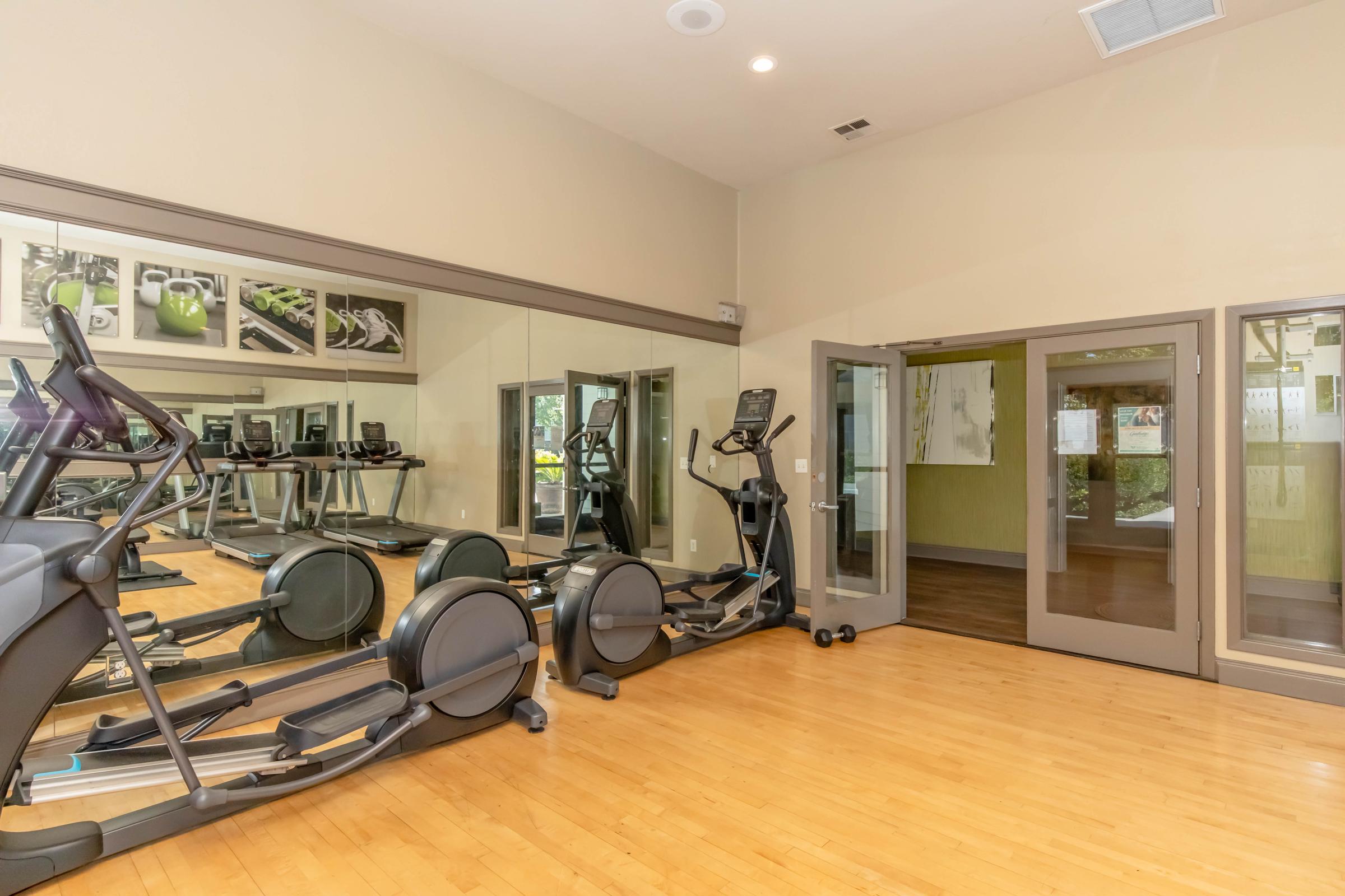 A spacious gym interior featuring mirrored walls, two ellipticals, and various fitness-related artwork. Natural light enters through large windows, illuminating the wooden floor. The atmosphere is clean and inviting, ideal for exercise and fitness activities.