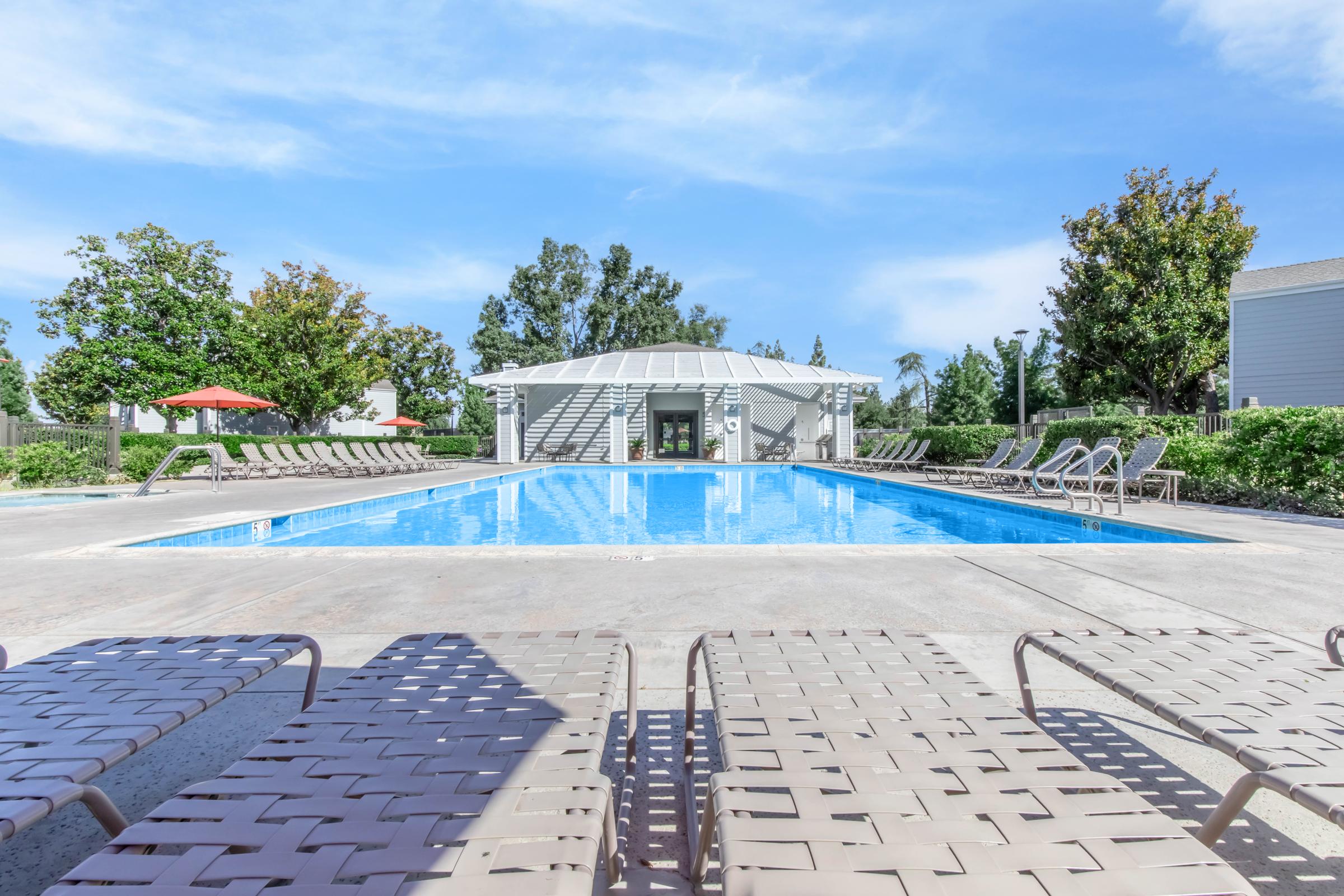 A spacious outdoor pool surrounded by lounge chairs on a sunny day. A covered pavilion is visible in the background, along with trees and shrubs. The sky is clear and blue, creating a relaxing atmosphere perfect for leisure and swimming.
