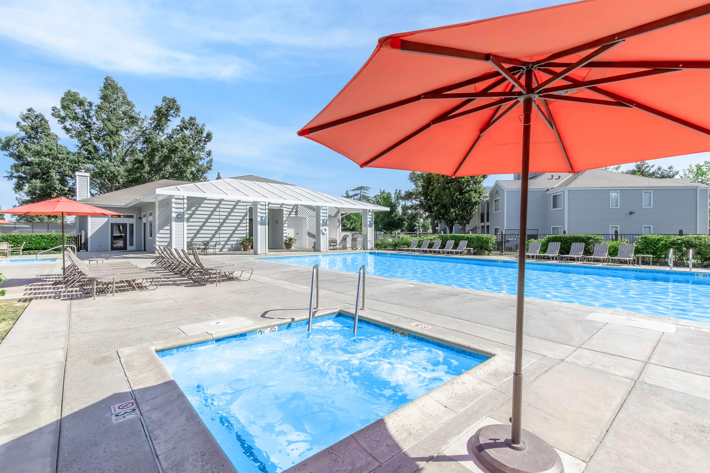 A sunny outdoor pool area featuring a large swimming pool, a hot tub, and several lounge chairs. Bright orange umbrellas provide shade, and a poolside building can be seen in the background. Lush green landscaping surrounds the area, creating a relaxing atmosphere.