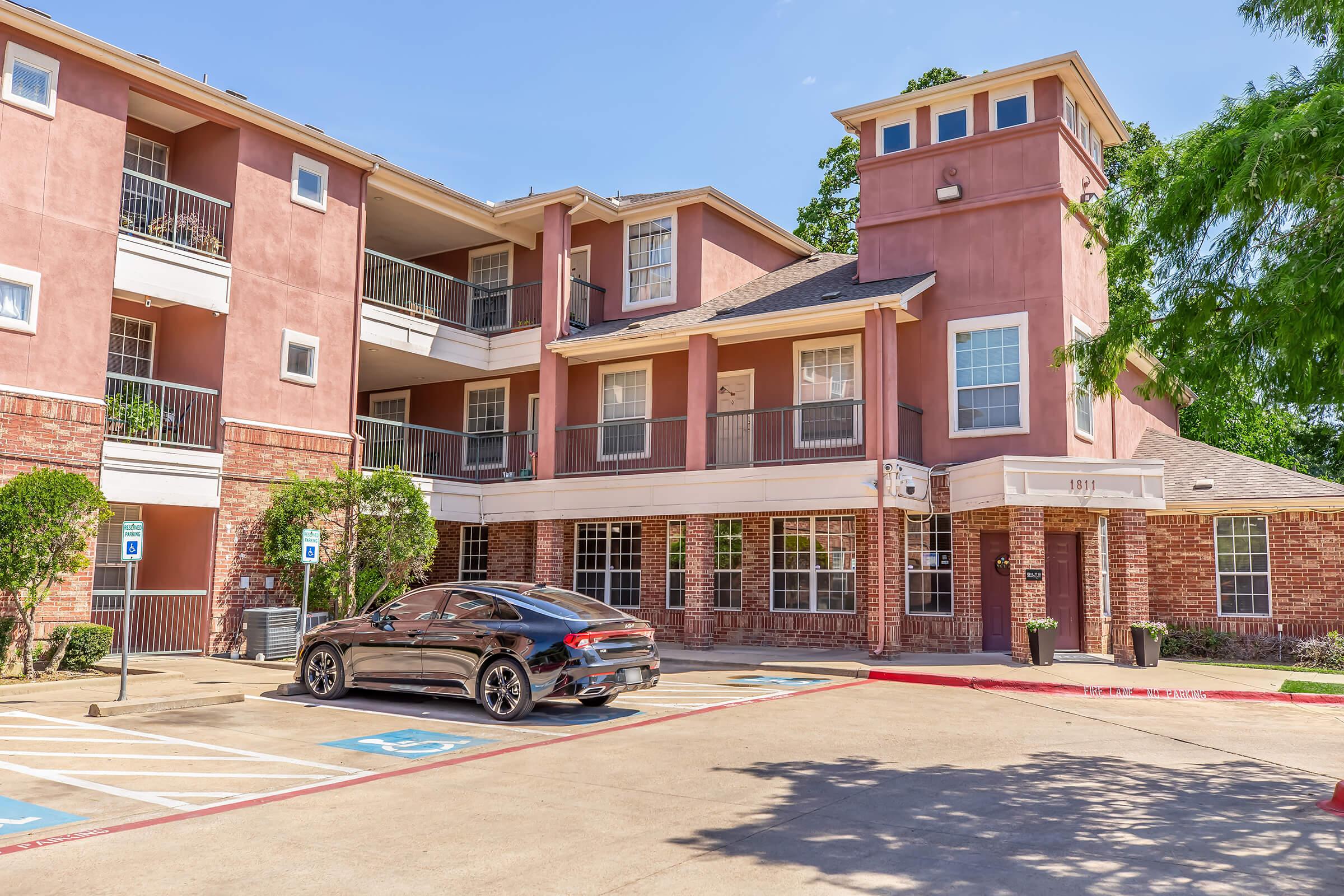 Exterior view of a multi-story apartment building featuring a mix of brick and stucco architecture. A parked black car is in the foreground, and there are marked parking spaces. The building has balconies and various windows, with well-maintained landscaping visible around the entrance.