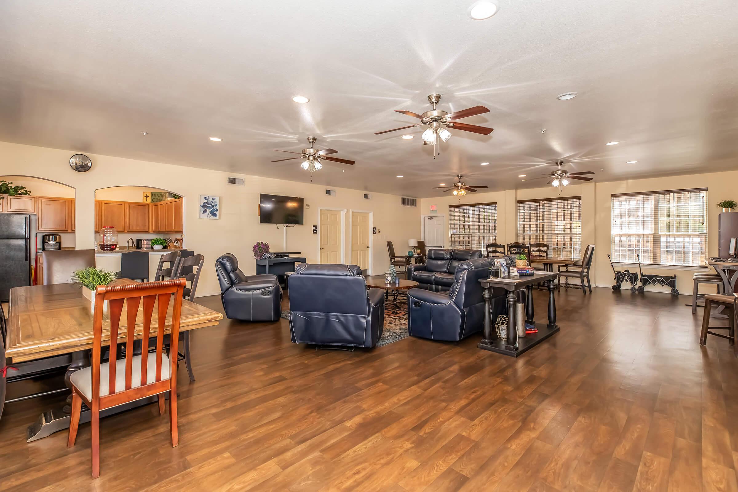 A spacious, well-lit communal living area featuring comfortable seating, a large dining table, and modern appliances. Ceiling fans and large windows enhance the atmosphere, which is suitable for socializing or relaxing. The flooring is wood-like, contributing to a cozy and inviting environment.