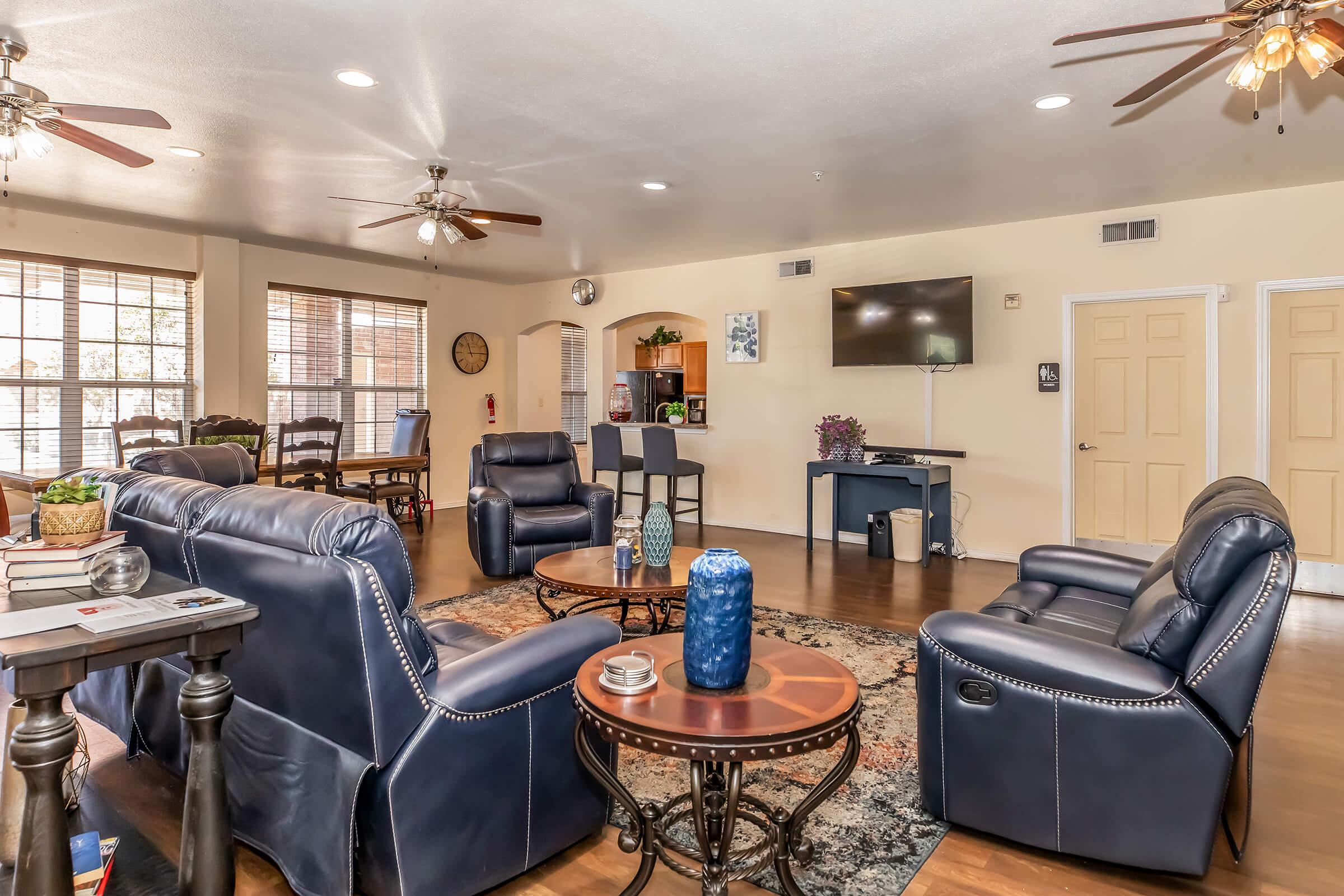 A cozy living room featuring comfortable leather seating, a round coffee table with a blue vase, and a wall-mounted TV. Large windows provide natural light, and the space includes a dining area in the background. The flooring is wooden, and the room has ceiling fans for ventilation.