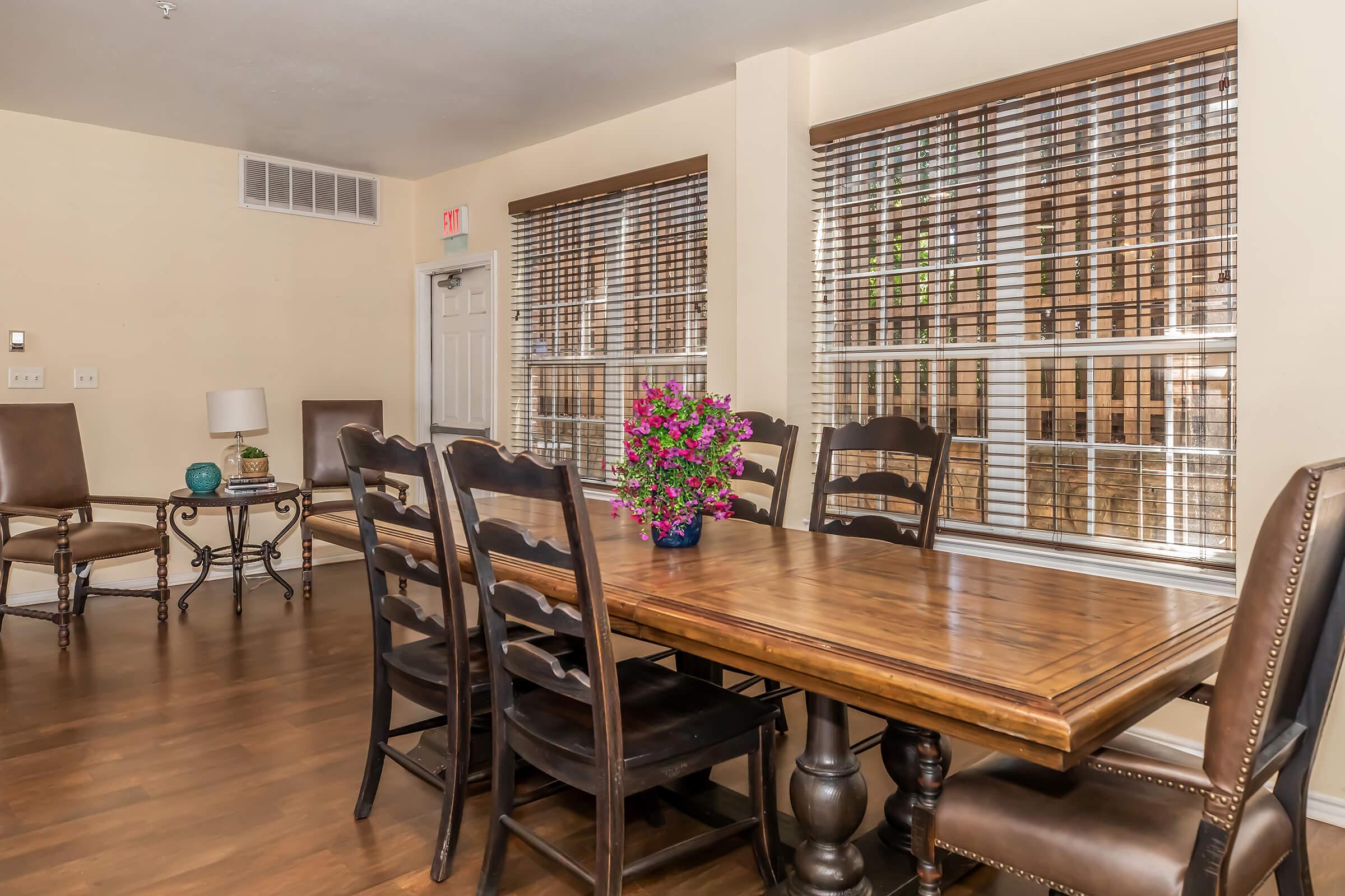 A well-lit dining area featuring a large wooden table surrounded by eight chairs. There is a vase of vibrant flowers on the table. The room has large windows with blinds, and a side table with a lamp and decorative items in the background, creating a warm and inviting atmosphere.