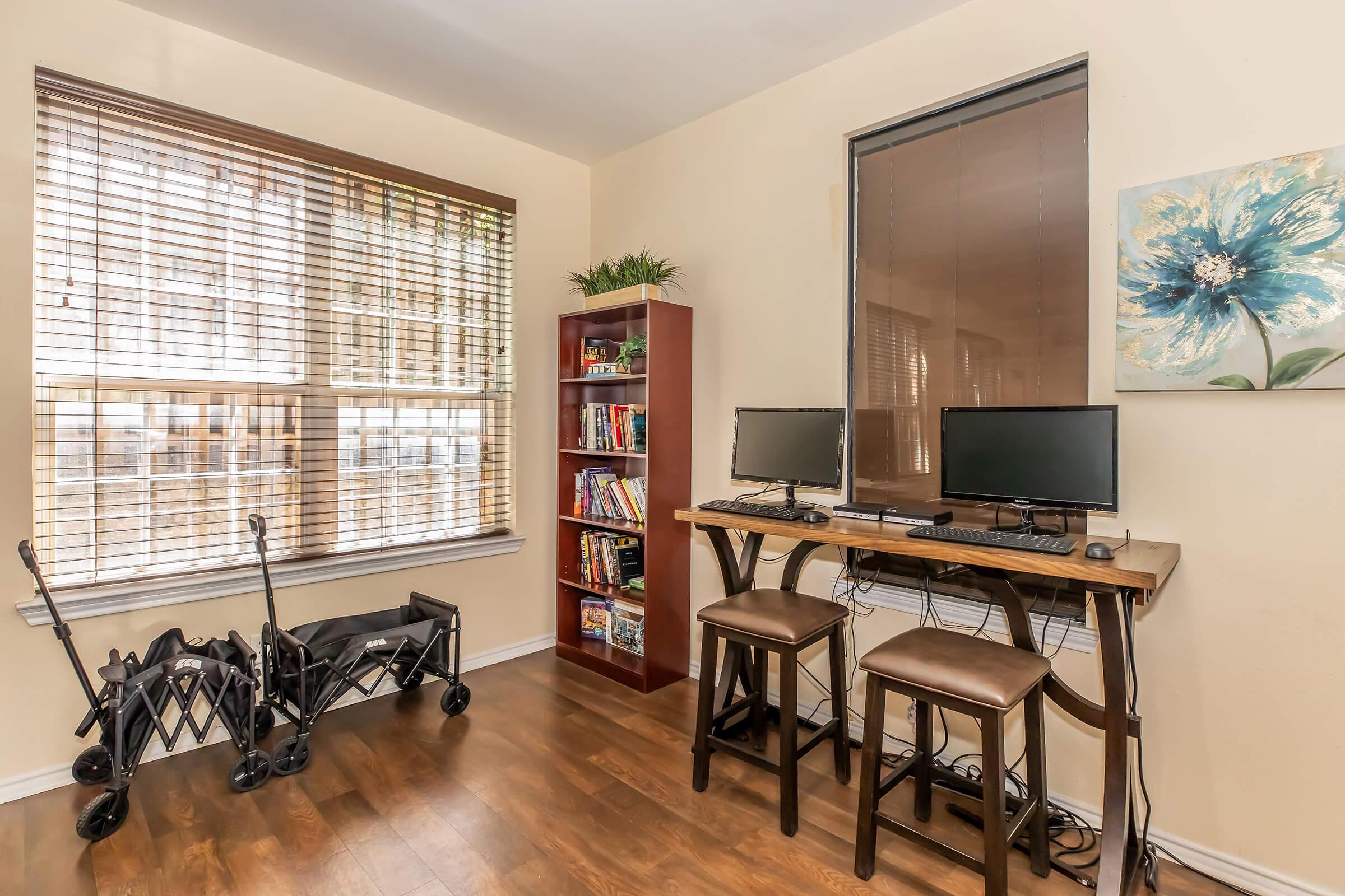 A cozy home office featuring two desktop computers on a wooden desk, with bar-style stools. A small bookshelf filled with books is nearby, and a stroller is positioned in the corner. Natural light filters through the window with blinds, creating a warm and inviting atmosphere.