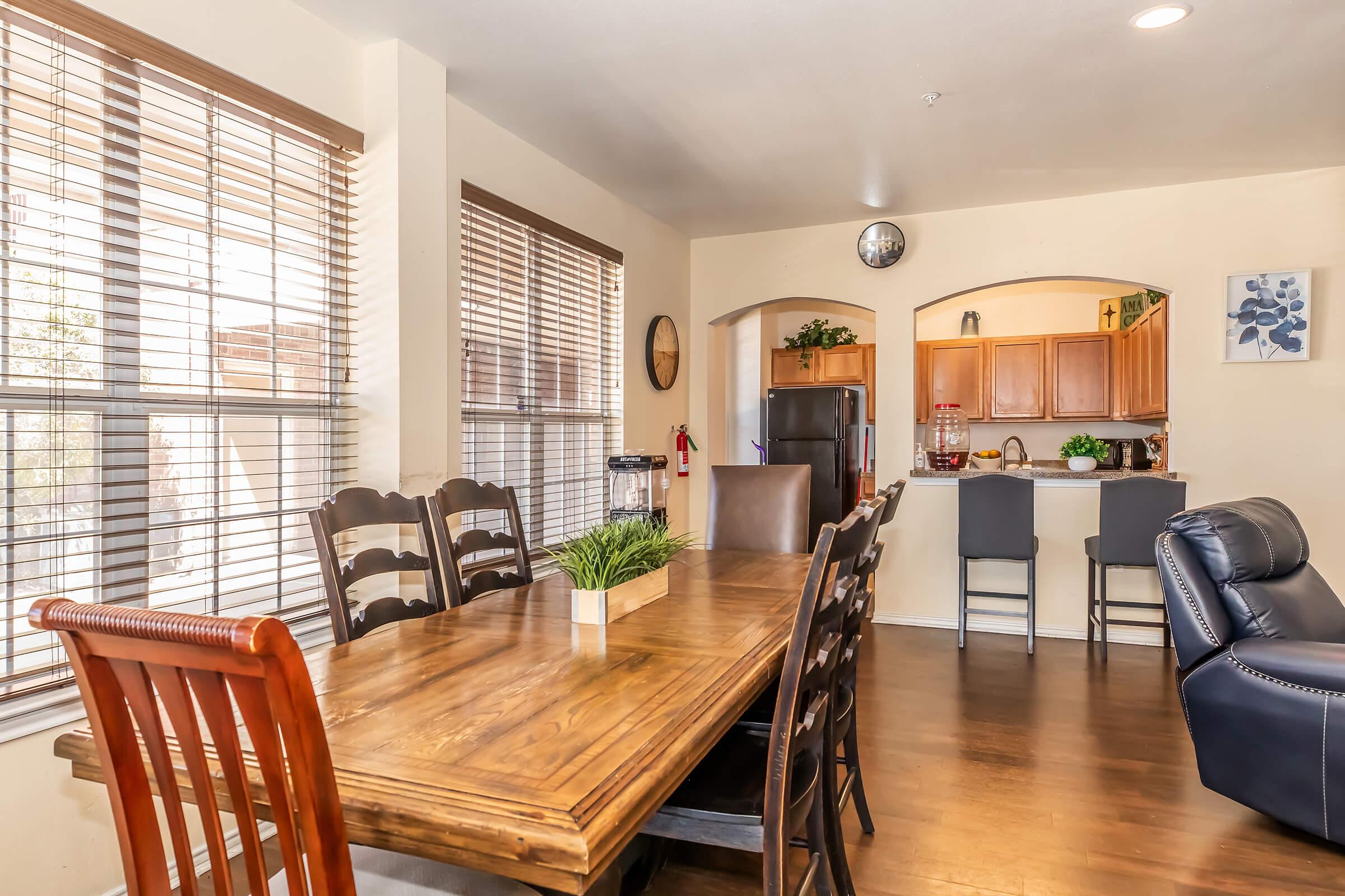 A spacious dining area featuring a wooden table with brown chairs, large windows with horizontal blinds, and a cozy kitchen in the background. The kitchen showcases wooden cabinets and modern appliances, while a potted plant adds a touch of greenery to the table.