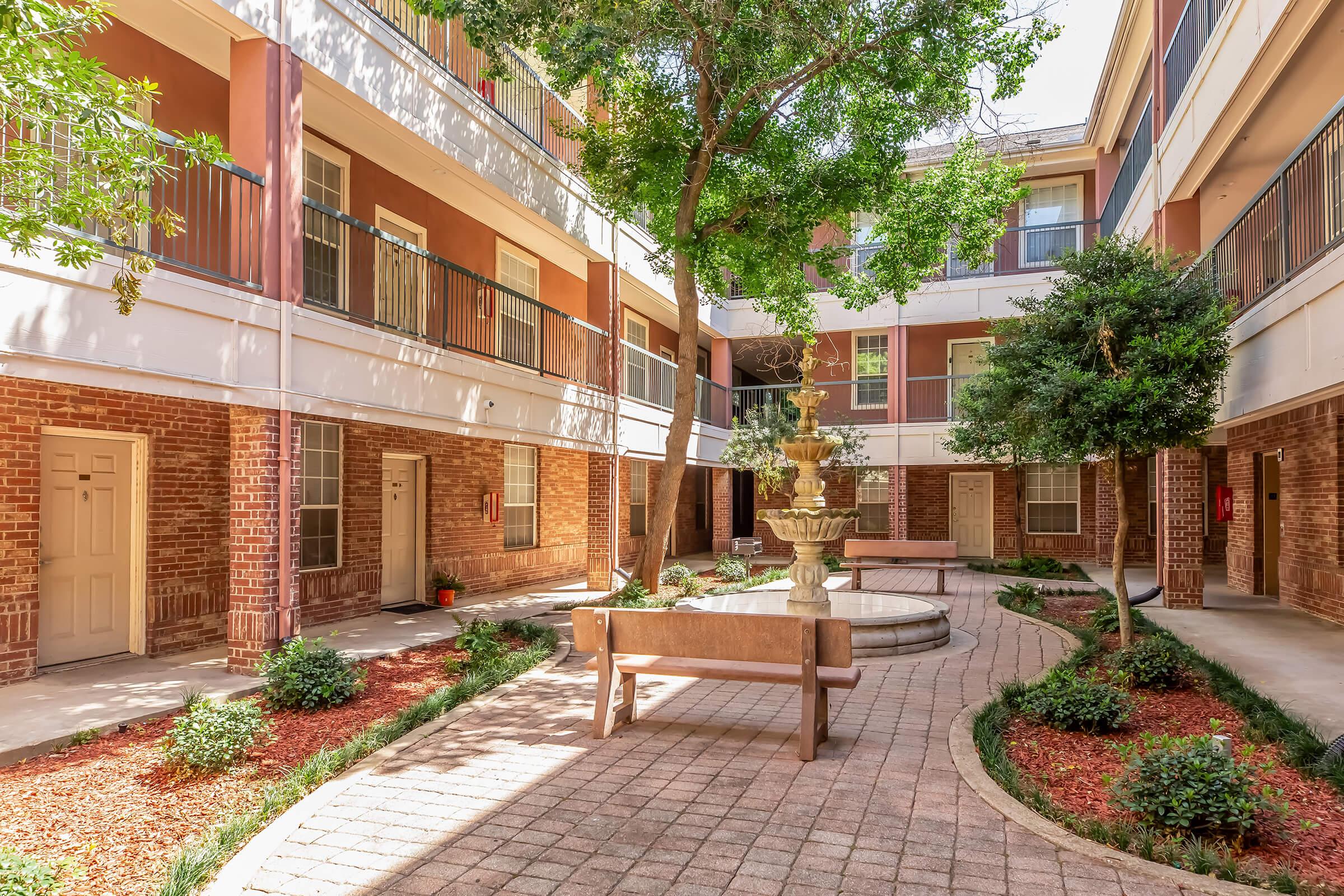 Outdoor courtyard of an apartment complex featuring stone pathways, landscaped greenery, and a decorative fountain surrounded by seating areas and balconies. The area is bright and inviting, with natural light filtering through the trees.