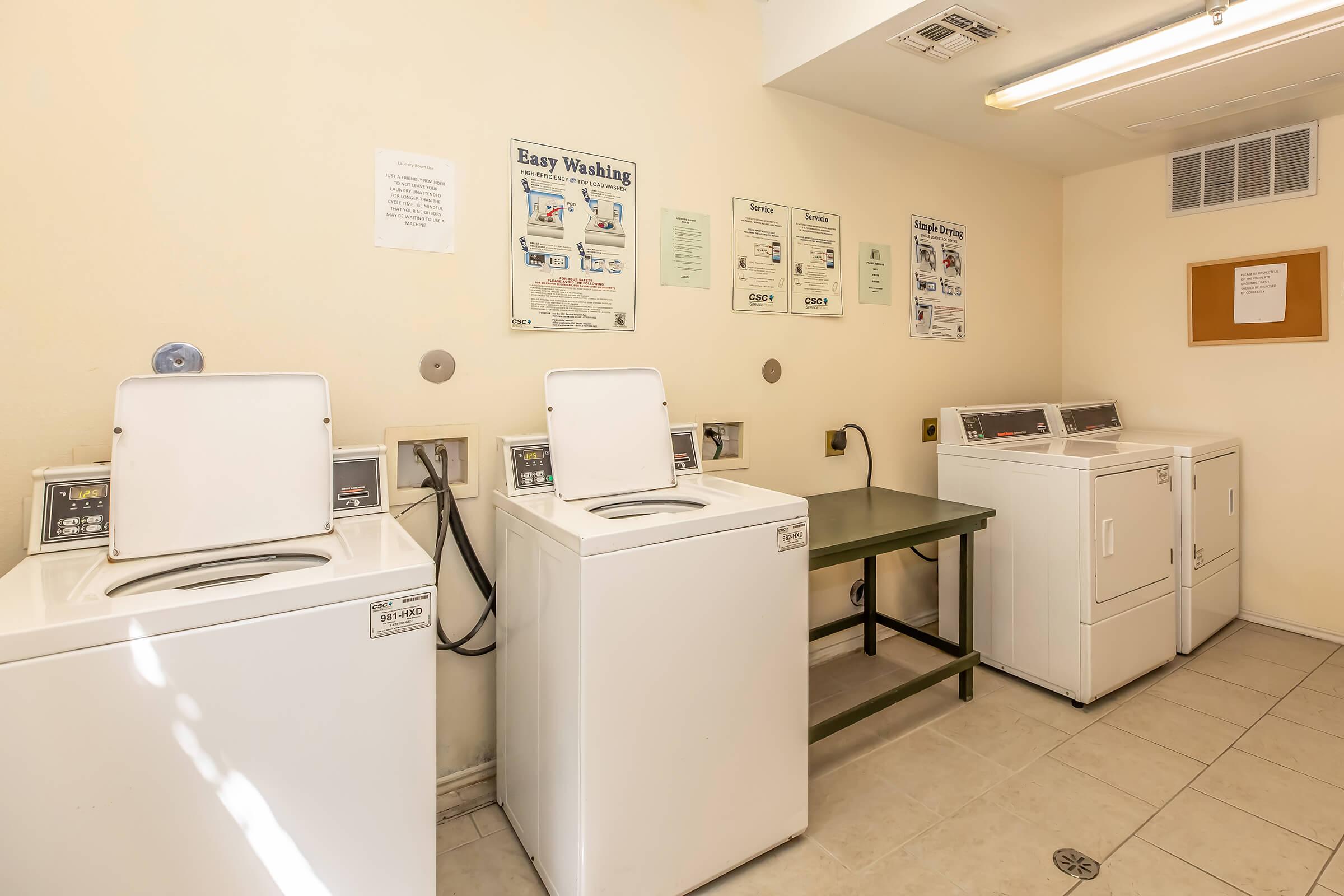 Laundry room featuring three white washing machines and a white dryer, all lined against a wall. A green table is positioned between the machines. Informative posters are displayed on the wall, detailing washing instructions and services. The space is well-lit and has tiled flooring.