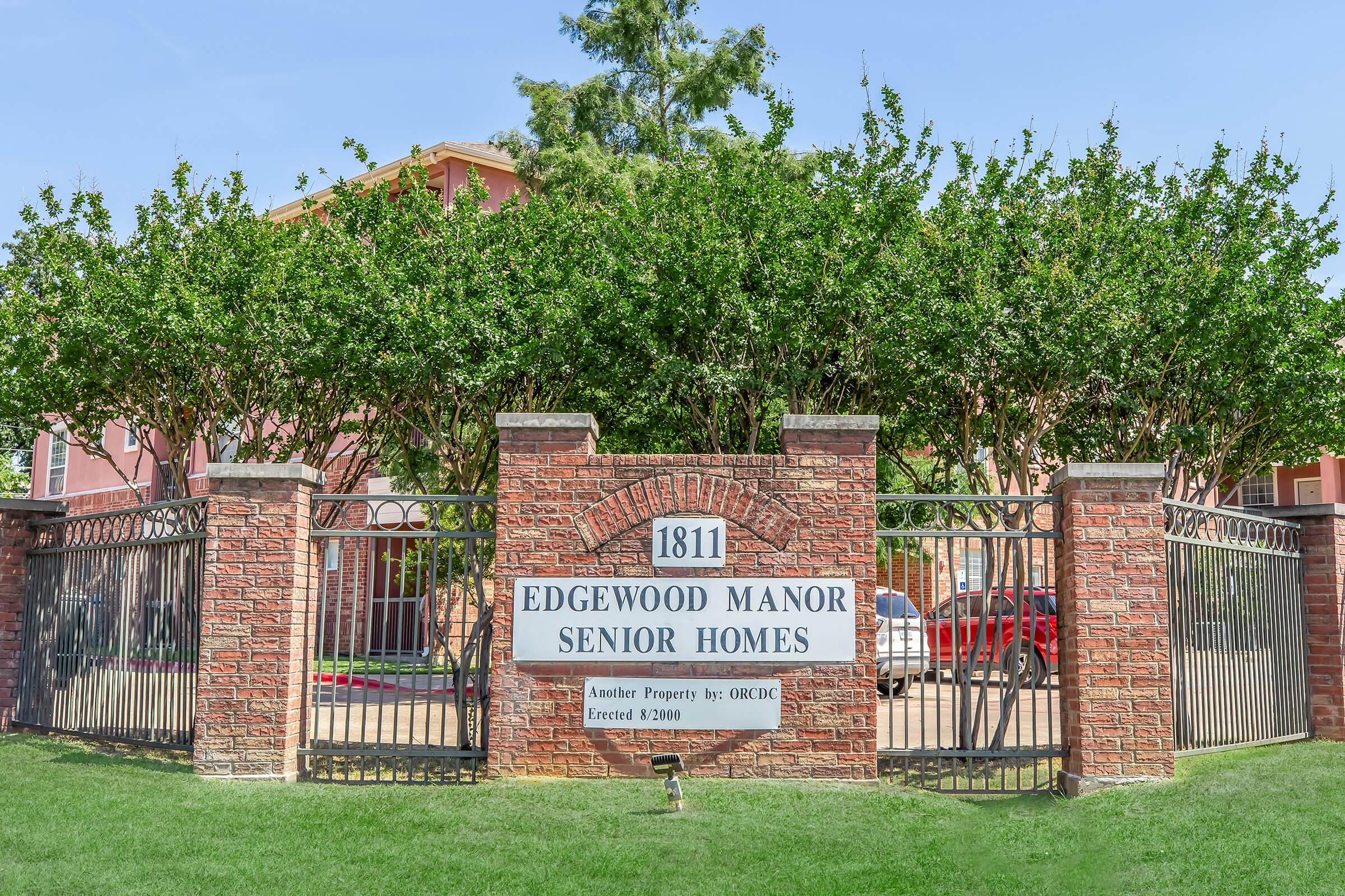Sign for Edgewood Manor Senior Homes, featuring a brick entrance with iron gates. The sign includes the address "1811" and specifies that it was established in 2000. Surrounding the entrance, there are trees and a well-maintained lawn.