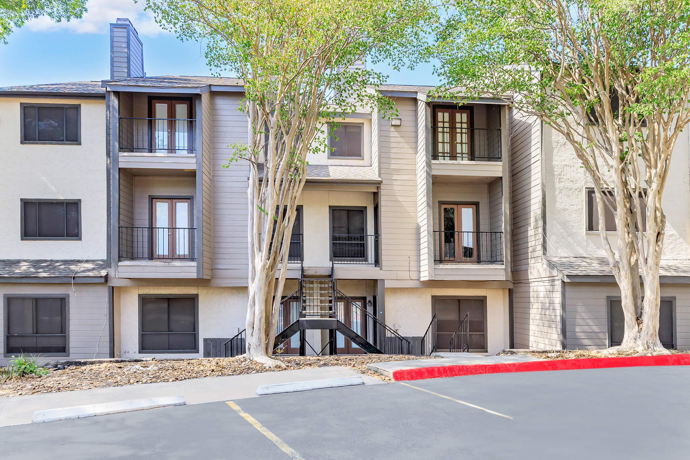 Three-story apartment building with a light gray exterior, featuring balconies on the second and third floors. Tall trees are planted in front, and a black staircase leads to the entrances. A parking lot in the foreground has a red curb. Clear blue sky visible above.