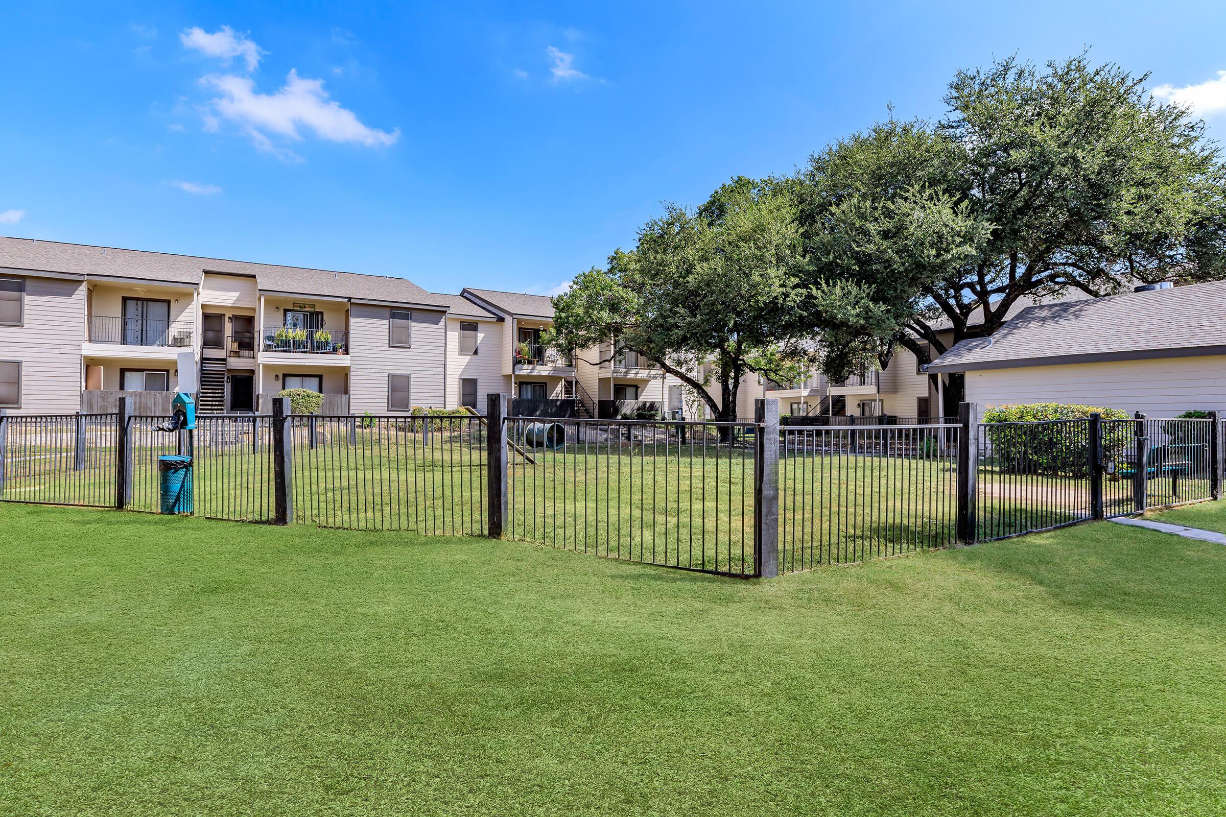 A well-maintained green lawn enclosed by a black metal fence, with several trees providing shade. In the background, there are two multi-story apartment buildings with balconies. The sky is bright blue with a few clouds, creating a pleasant outdoor scene.