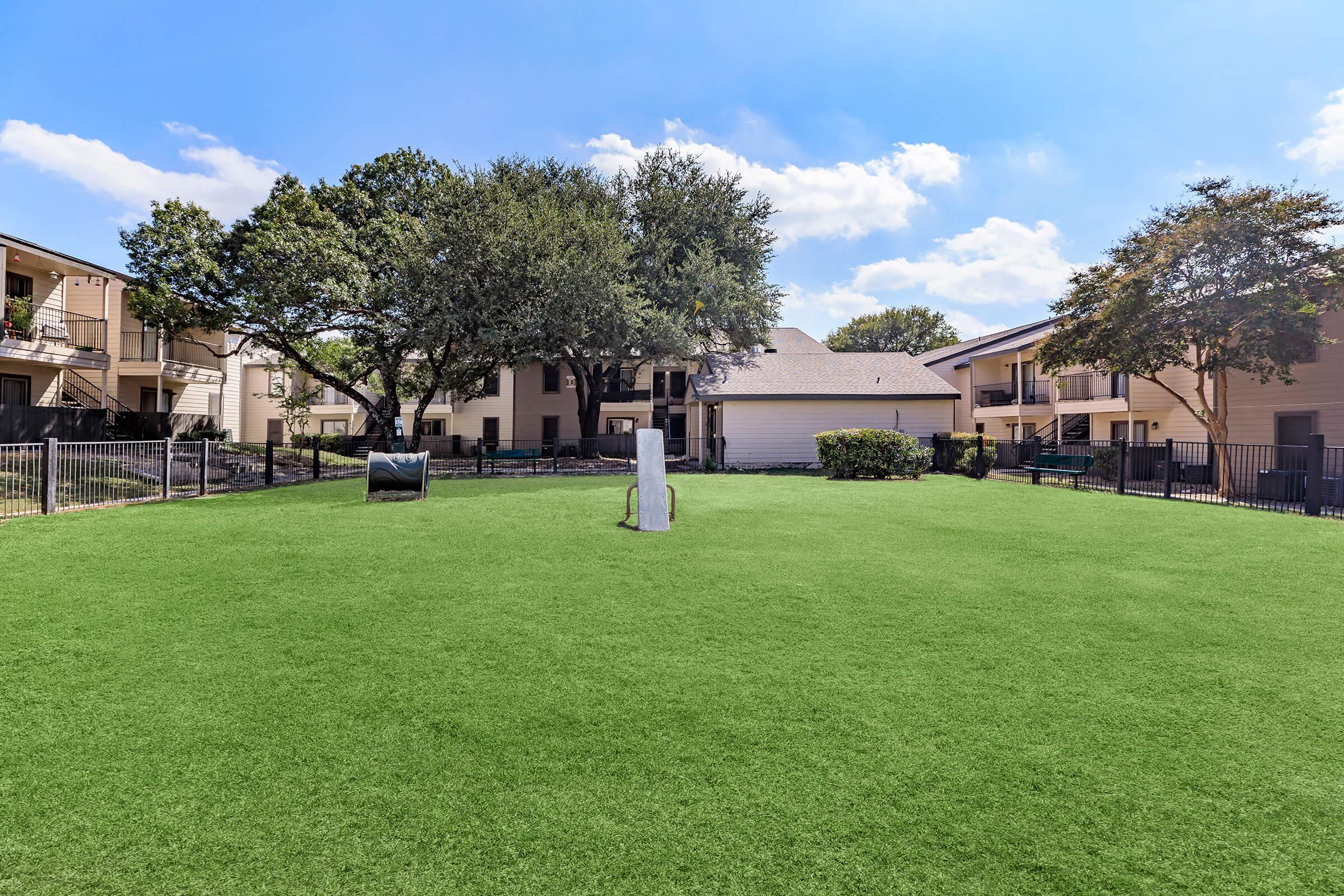 A well-maintained grassy area with artificial turf, featuring trees on the edges and an apartment complex in the background. The space includes a small bush and a dog waste station, creating an inviting outdoor environment. The sky is clear with a few clouds, suggesting a sunny day.
