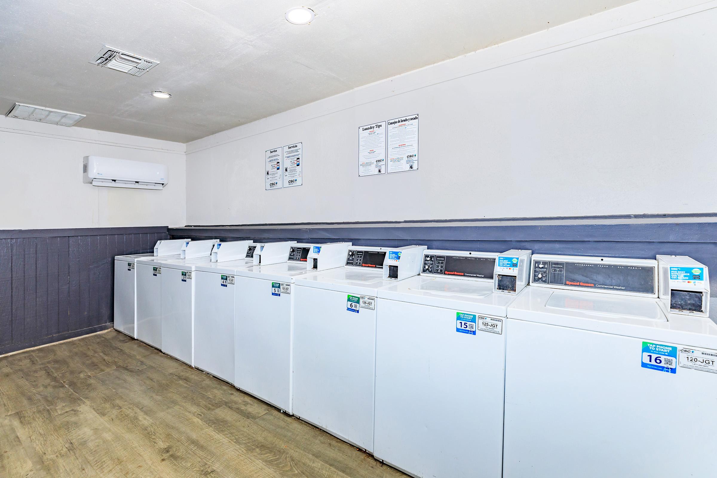 A clean and organized laundry room featuring several white washing machines in a row. The walls are painted light colors, and there's an air conditioning unit on the wall. Informational posters are displayed above the machines. The floor is made of wooden planks, creating a welcoming atmosphere.