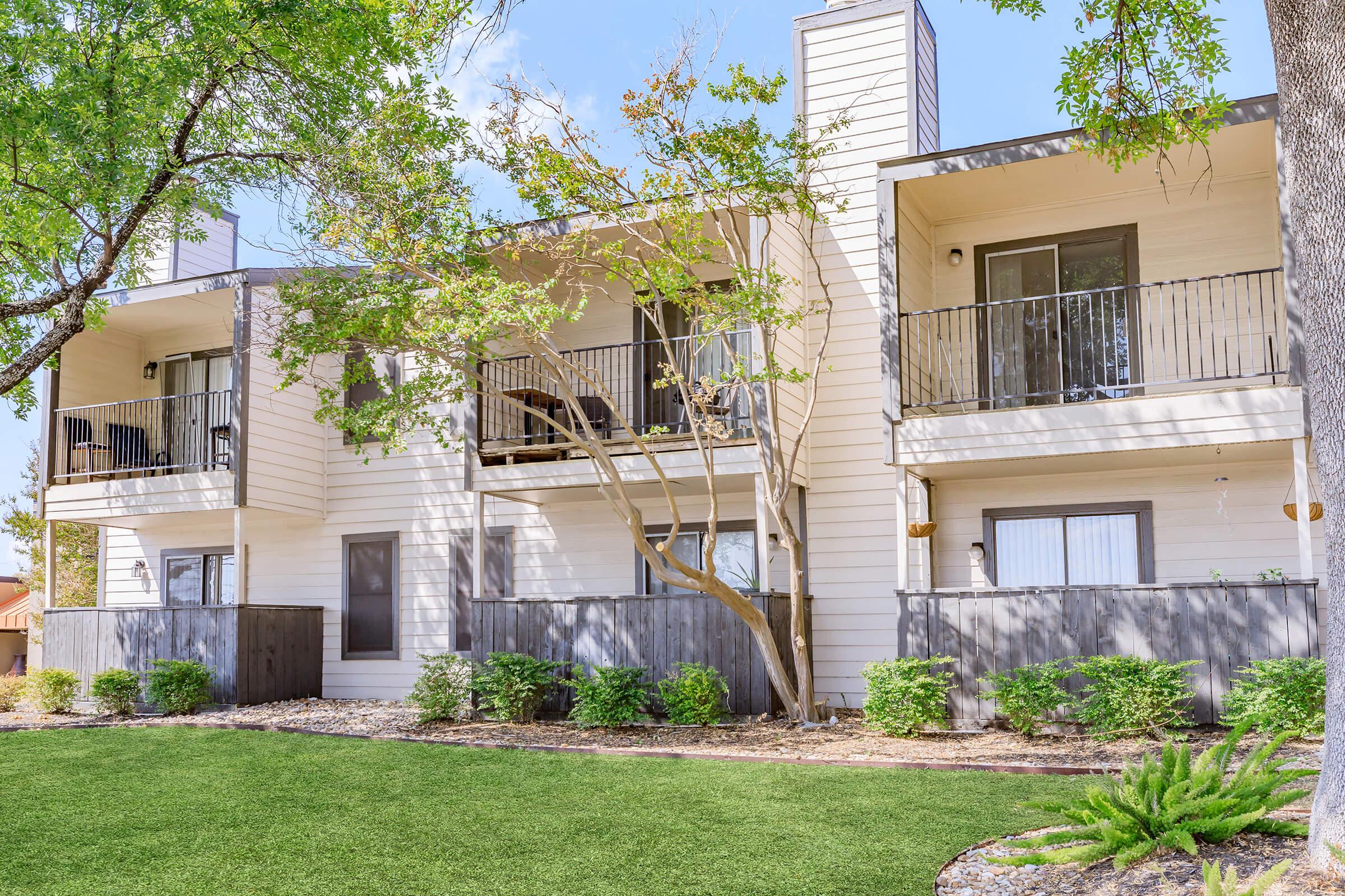 A multi-unit residential building with three balconies, surrounded by trees and landscaped greenery. The building features light-colored exterior walls and a well-maintained lawn in front, creating a pleasant, inviting atmosphere.
