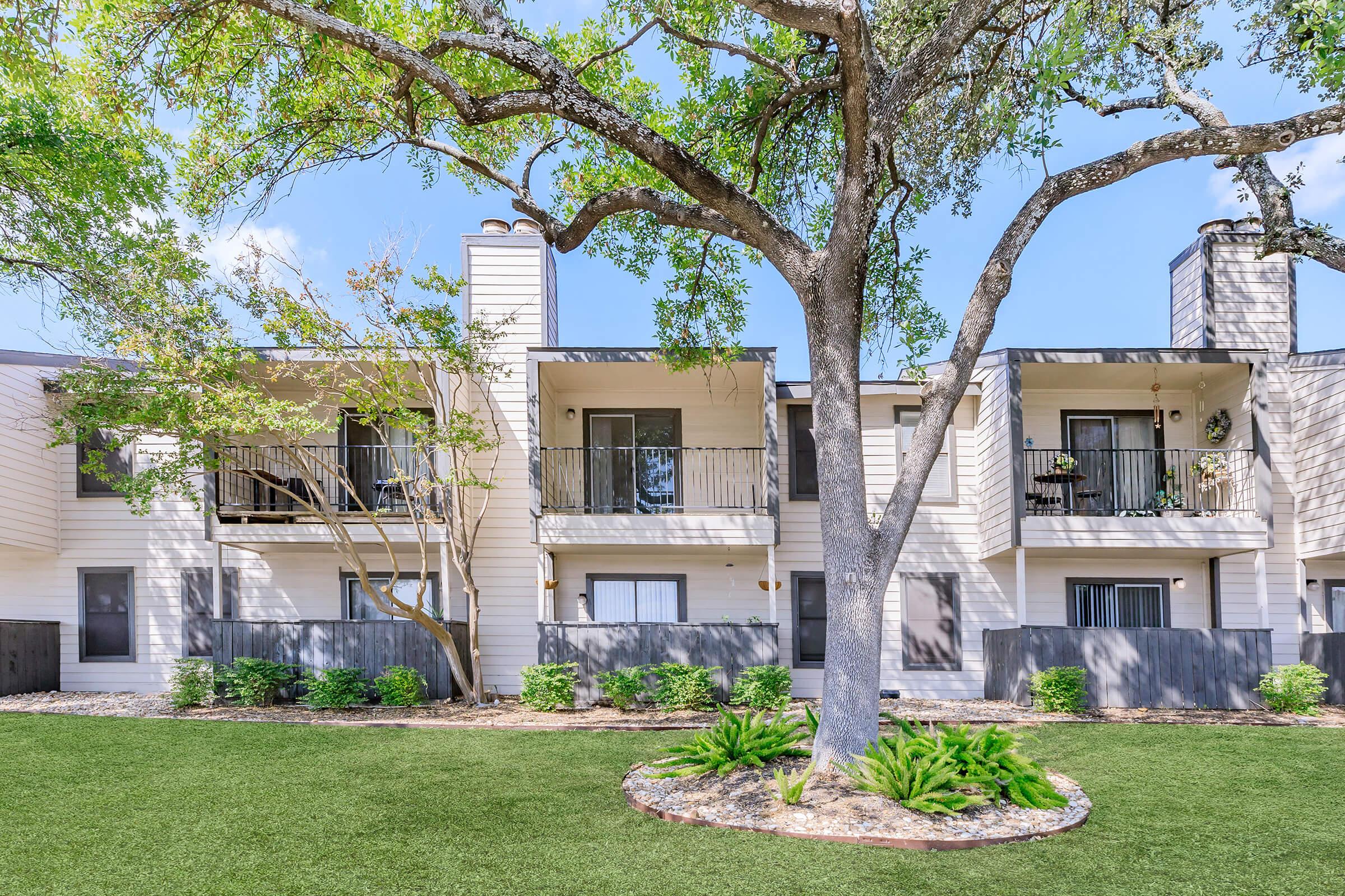 Modern apartment complex featuring two-story buildings with balconies. A large tree and landscaped greenery are visible in the foreground, creating a welcoming outdoor space. The sky is clear and blue, enhancing the inviting atmosphere of the residential area.