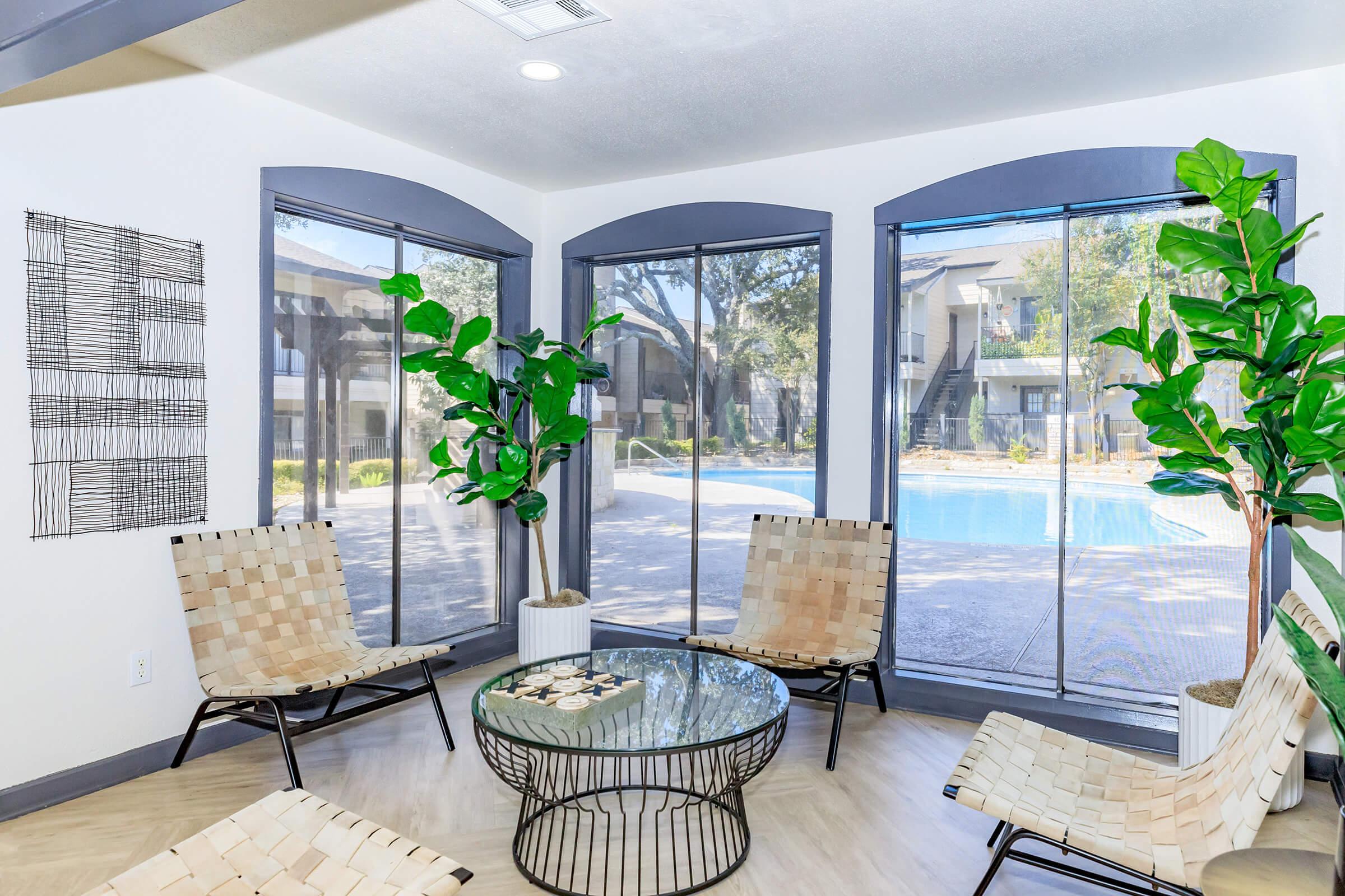 Bright, modern lounge area with large windows showcasing a pool view. The space features minimalist decor, including woven chairs and a round glass table. Two potted plants add a touch of greenery, enhancing the inviting atmosphere. A piece of abstract wall art is displayed on the left.