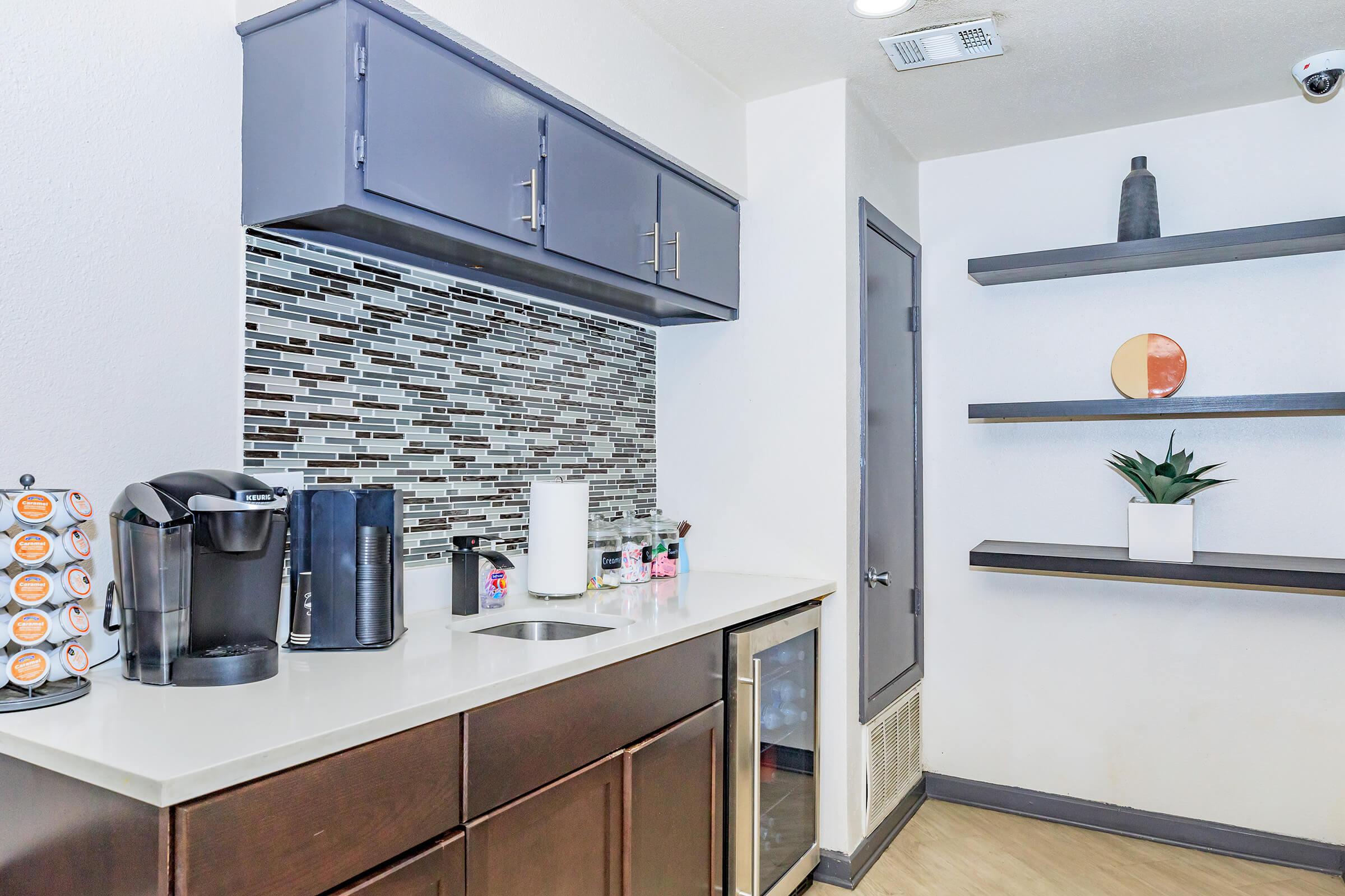 A modern kitchen area featuring dark wood cabinets, a coffee maker, a small fridge, and a decorative wall with a mosaic tile backsplash. Open shelving with minimalist decor items is visible, along with a white paper towel holder and a potted plant on the counter.