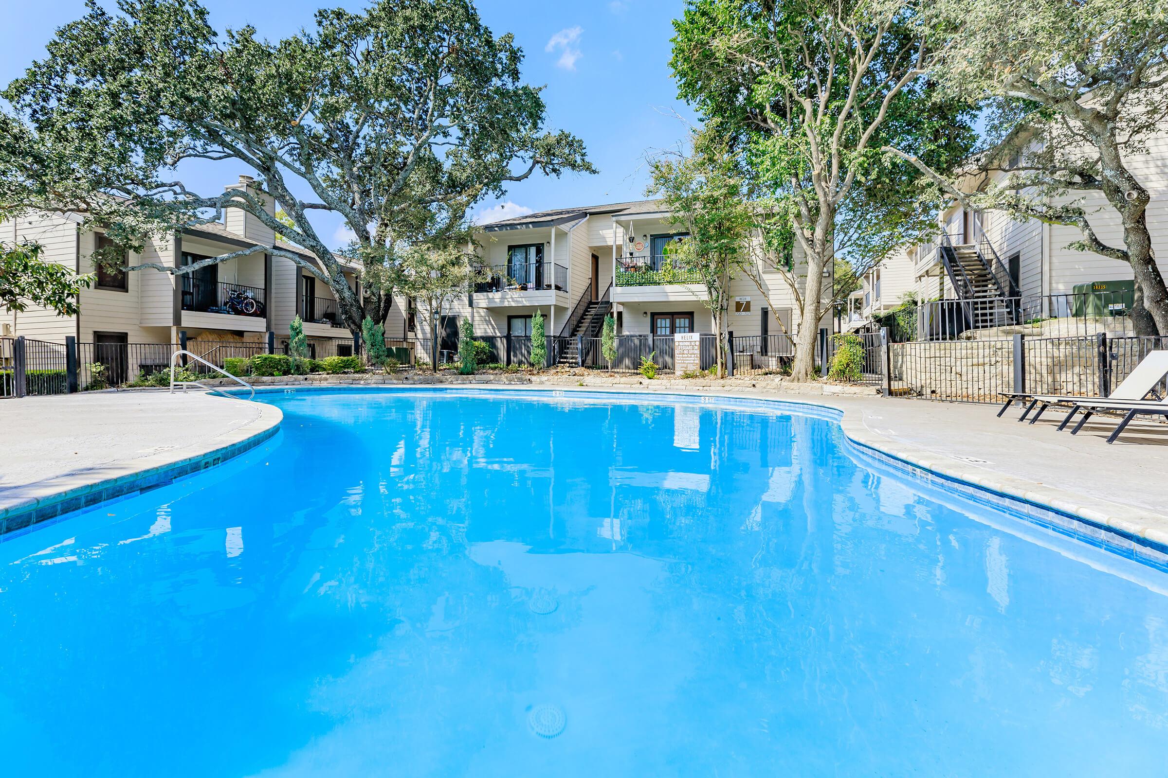 Clear blue swimming pool surrounded by lush trees and landscaped grounds. Apartment buildings with balconies are visible in the background, and there are lounge chairs near the pool. The scene is sunny and inviting, suggesting a peaceful outdoor area for relaxation.