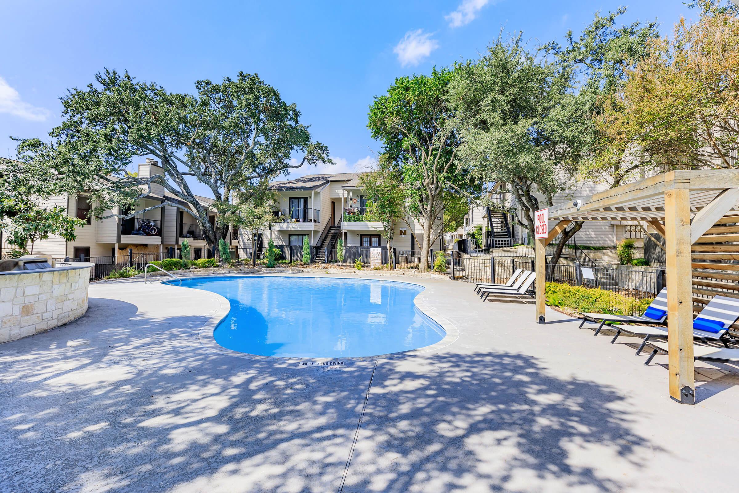 A sunny apartment complex pool area with a blue swimming pool surrounded by lounge chairs. Lush green trees and well-maintained landscaping create a relaxing atmosphere. The sky is clear with a few clouds, enhancing the bright and inviting ambiance of the space.