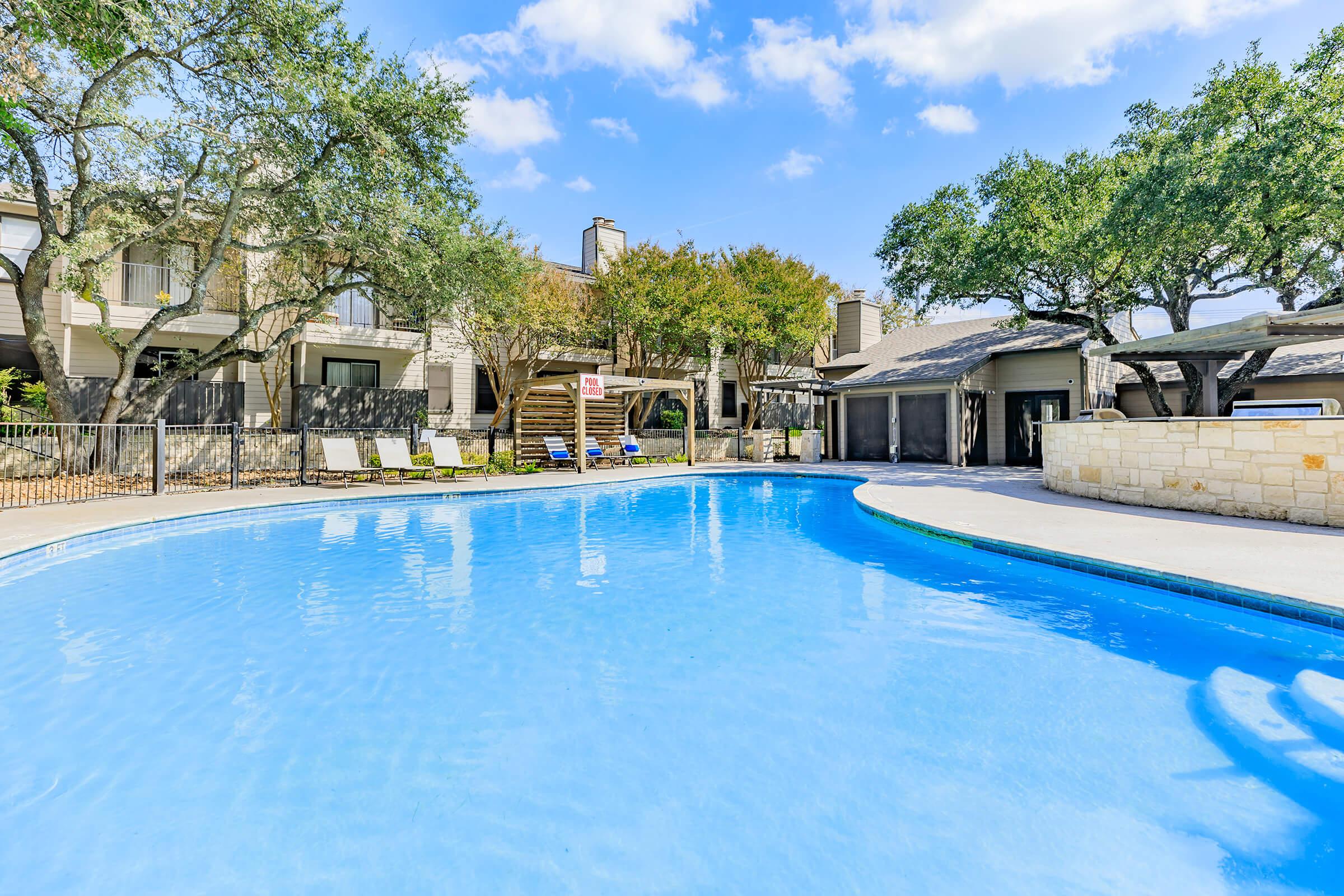 A clear blue swimming pool surrounded by a spacious patio area. There are several lounge chairs near the pool, trees providing shade, and two adjacent buildings in the background. The scene is bright and inviting, suggesting a relaxing outdoor space.