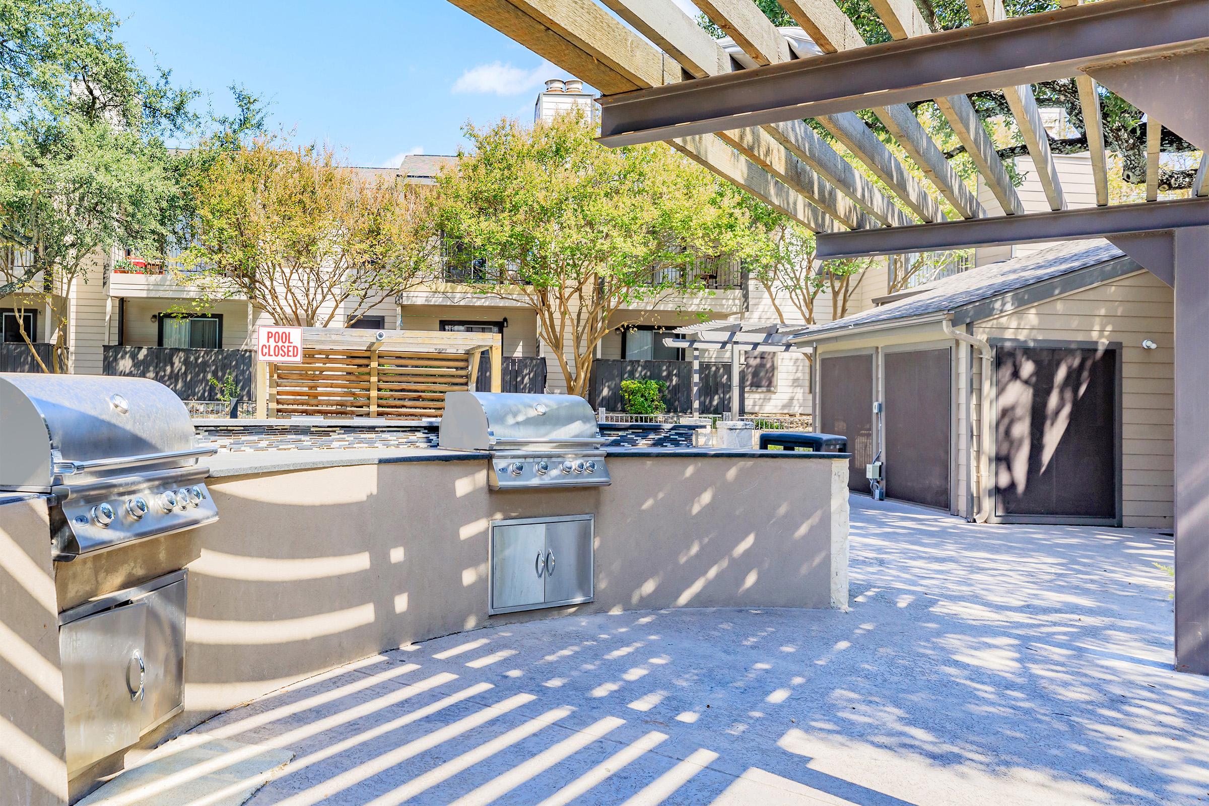 Outdoor kitchen area featuring several stainless steel grills under a wooden pergola. The space is well-lit with sunlight casting shadows on the ground. In the background, there are trees and a sign indicating the pool is closed. Two garages are visible on the right side.