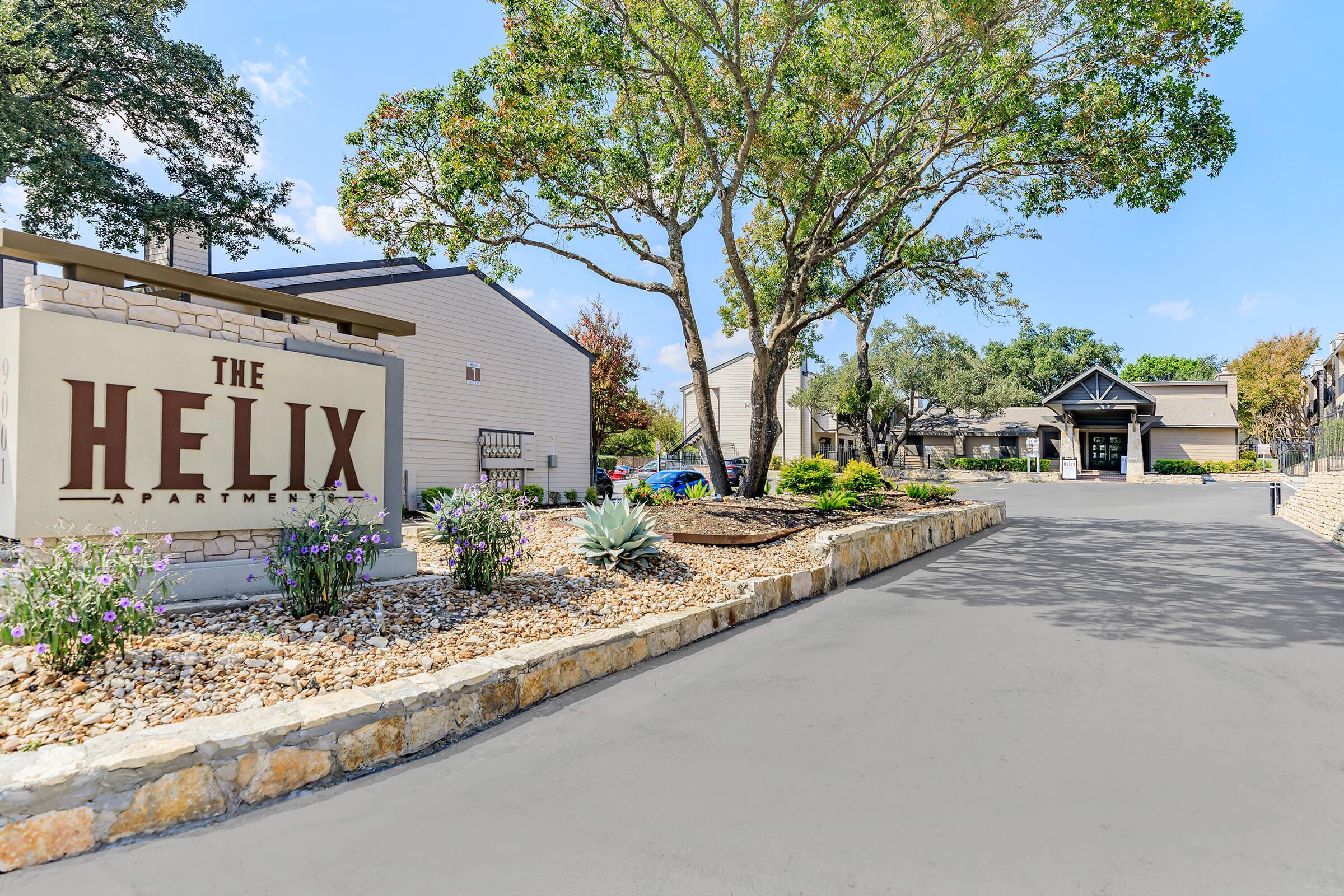 Sign for "The Helix Apartments" at the entrance, surrounded by landscaped greenery and a stone pathway. The scene features trees, shrubs, and colorful flowers, set against a clear blue sky. In the background, modern apartment buildings are partially visible.