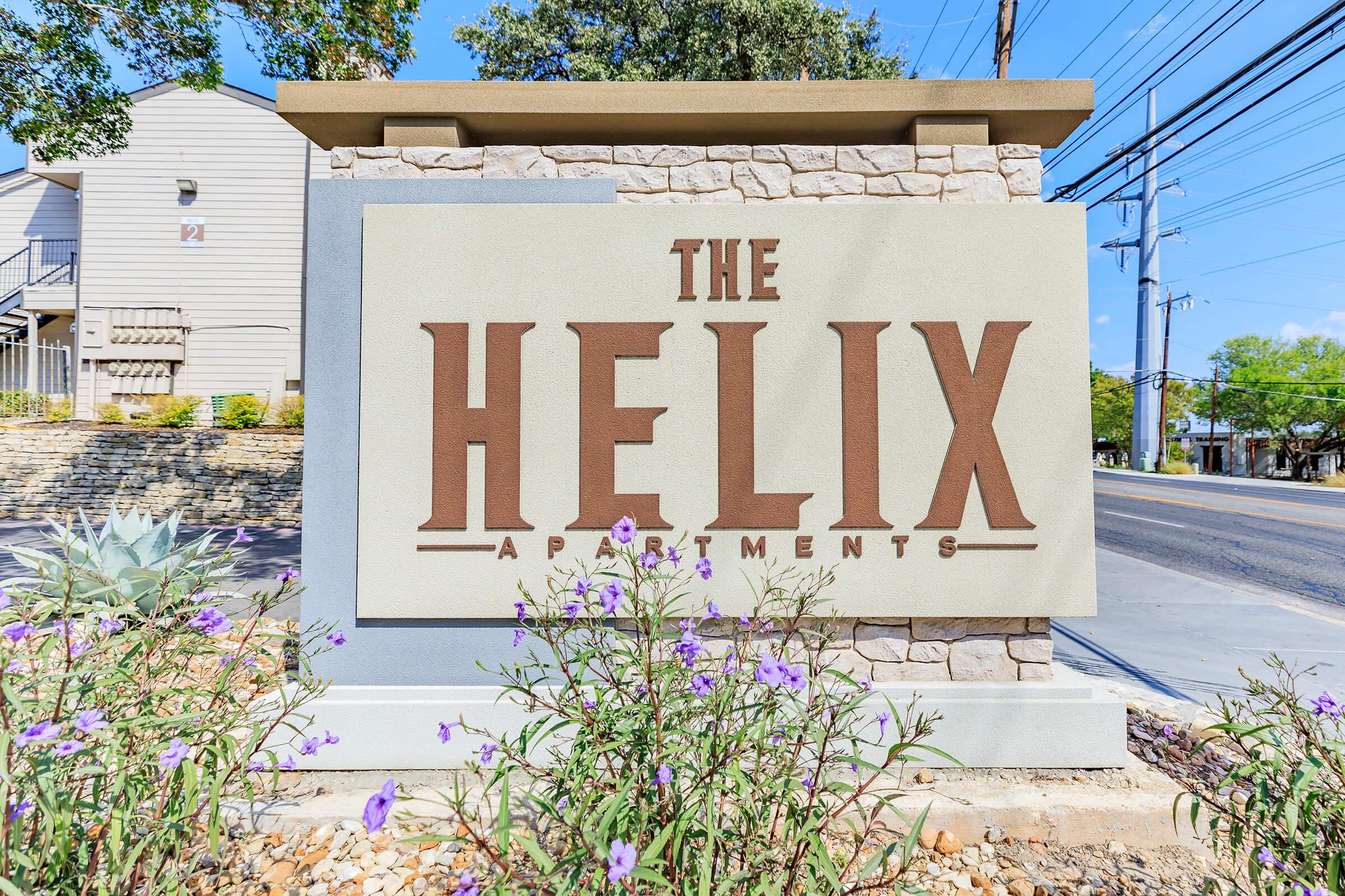 Sign for The Helix Apartments, featuring the name prominently in stylized lettering. The sign is set in a landscaped area with rocks and plants, and there are electrical poles and a blue sky in the background.
