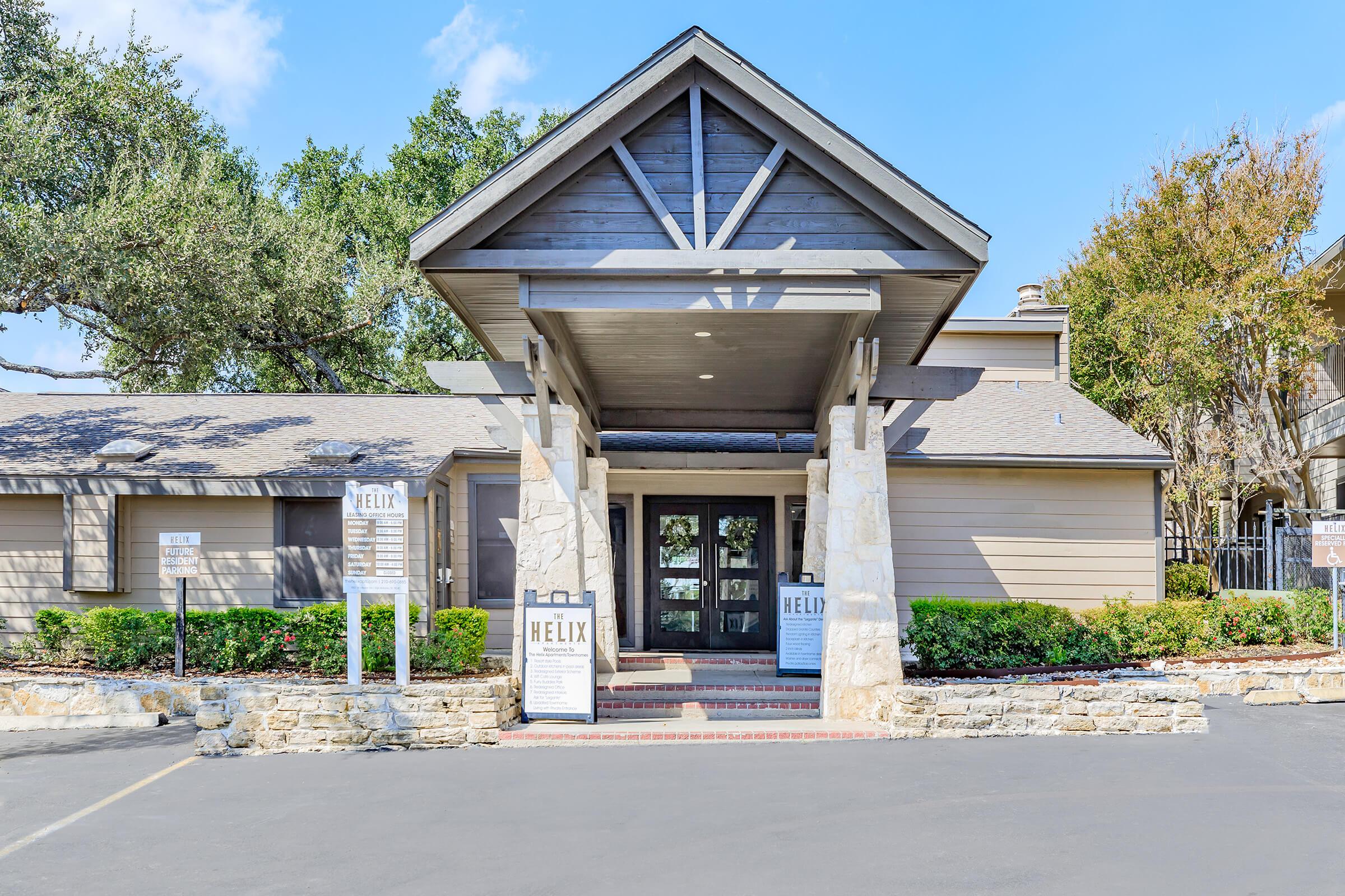 Front view of the Helix building, featuring a wooden entrance with a peaked roof and stone pillars. The entrance is flanked by signs indicating it is the Helix facility. Surrounding the building are well-maintained shrubs and trees under a clear blue sky. A paved driveway leads up to the entrance.